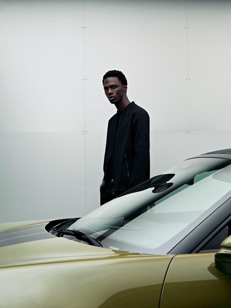 Homme en veste noire devant un mur blanc, au premier plan le pare-brise et le capot d’une Porsche dorée.