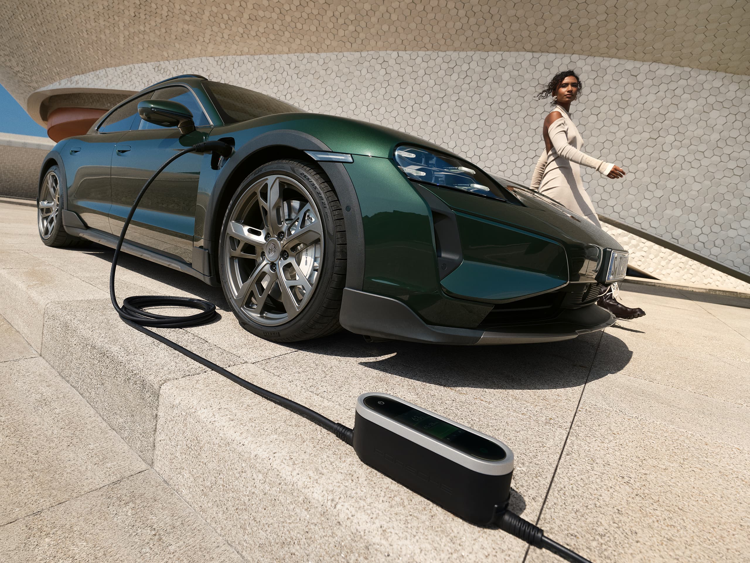 Green Porsche Taycan charging on a concrete platform, woman in beige dress walking by, modern architecture in background.