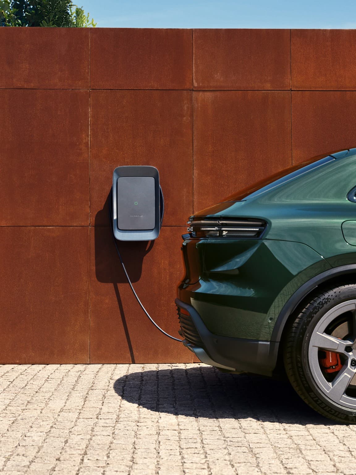 Grüner Porsche lädt an einer wandmontierten Station an einer rostfarbenen Wand, sonniger Tag, Kopfsteinpflaster.