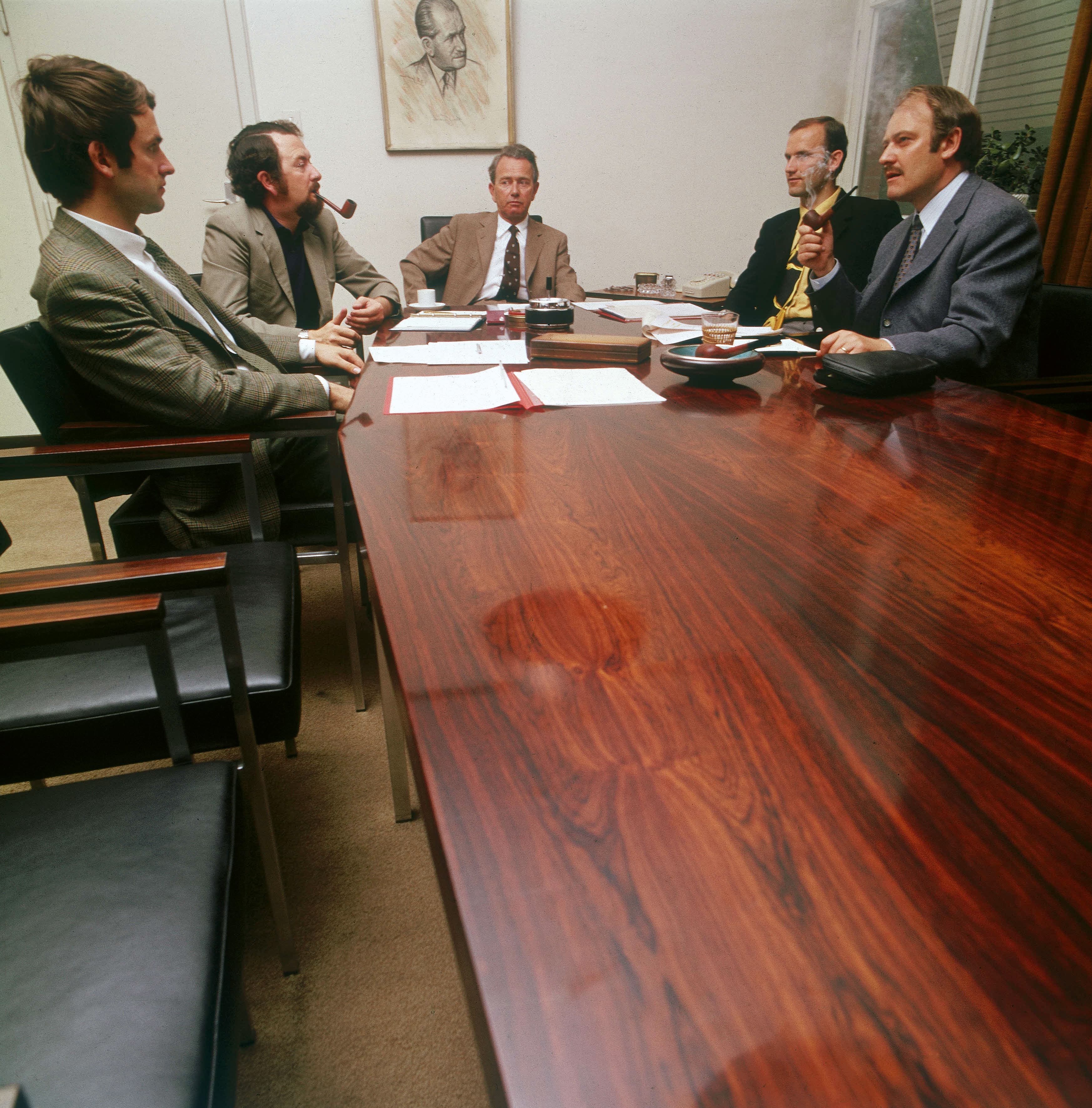 Five men in suits are sitting at a conference table with papers; two are smoking pipes, portrait on the wall.