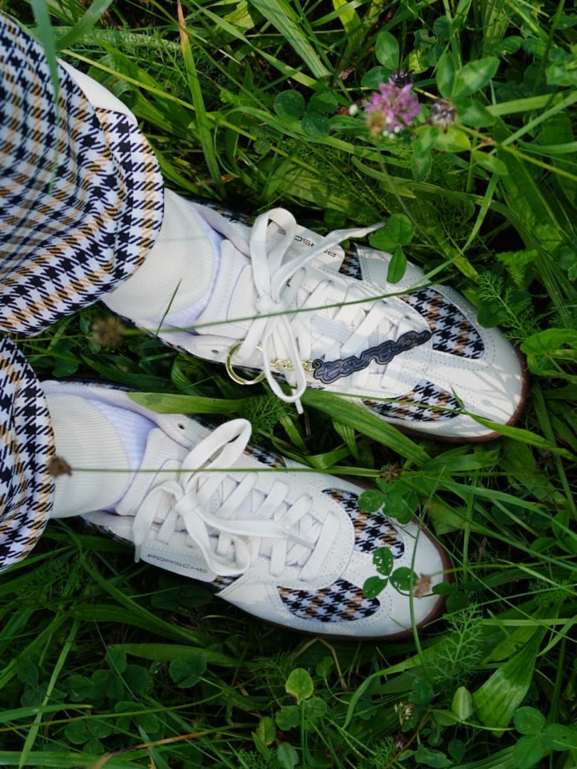 Person holding gray, black, and blue Porsche sneakers with striped design; wearing white sneakers on a tiled floor.