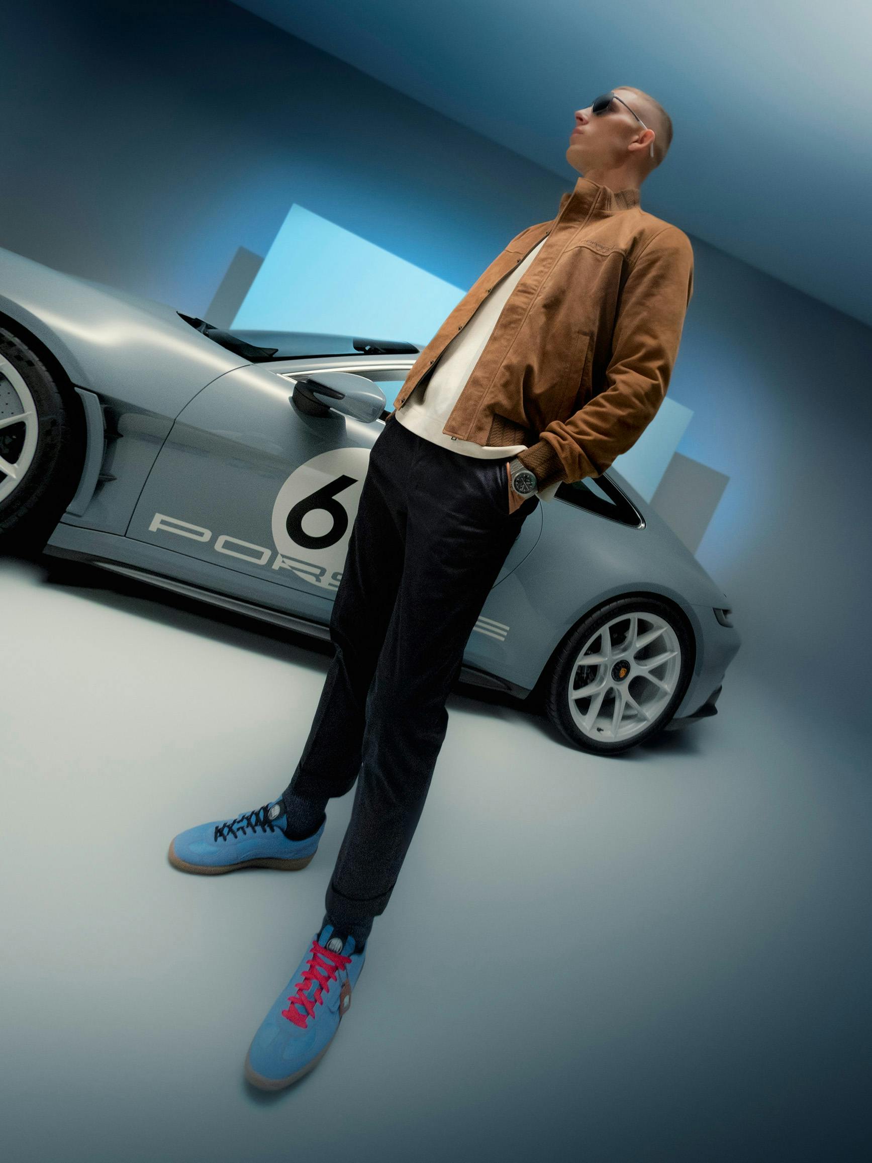 A man wearing a brown jacket, sunglasses and the new Puma sneakers in cooperation with Porsche can be seen from the side. A blue 911 Carrera S/T is cut off in the background.