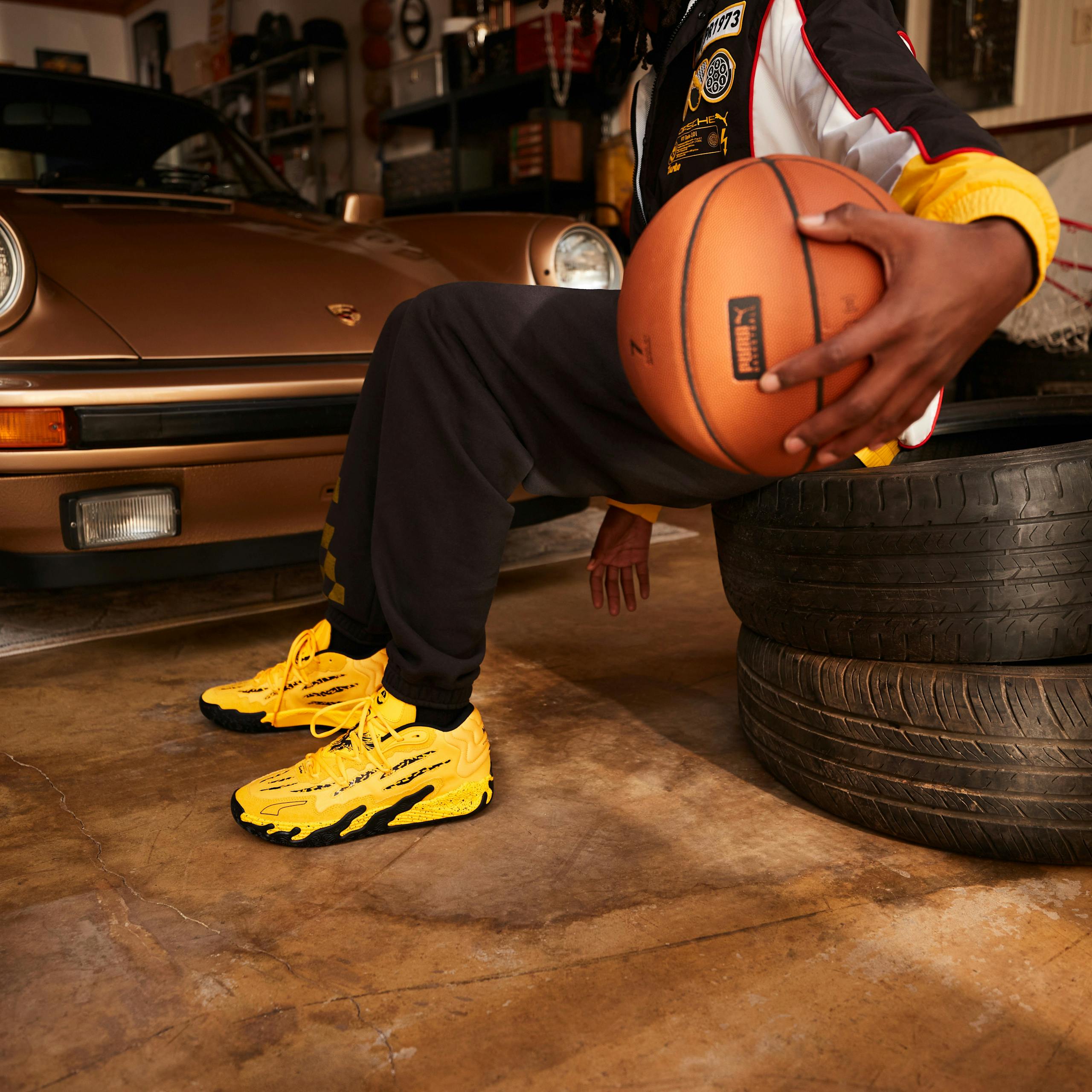 Porsche x Puma Basketball, image features man in garage dribbling basketball. He is sitting on a stack of tires with a Porsche 911 Turbo in the background. 