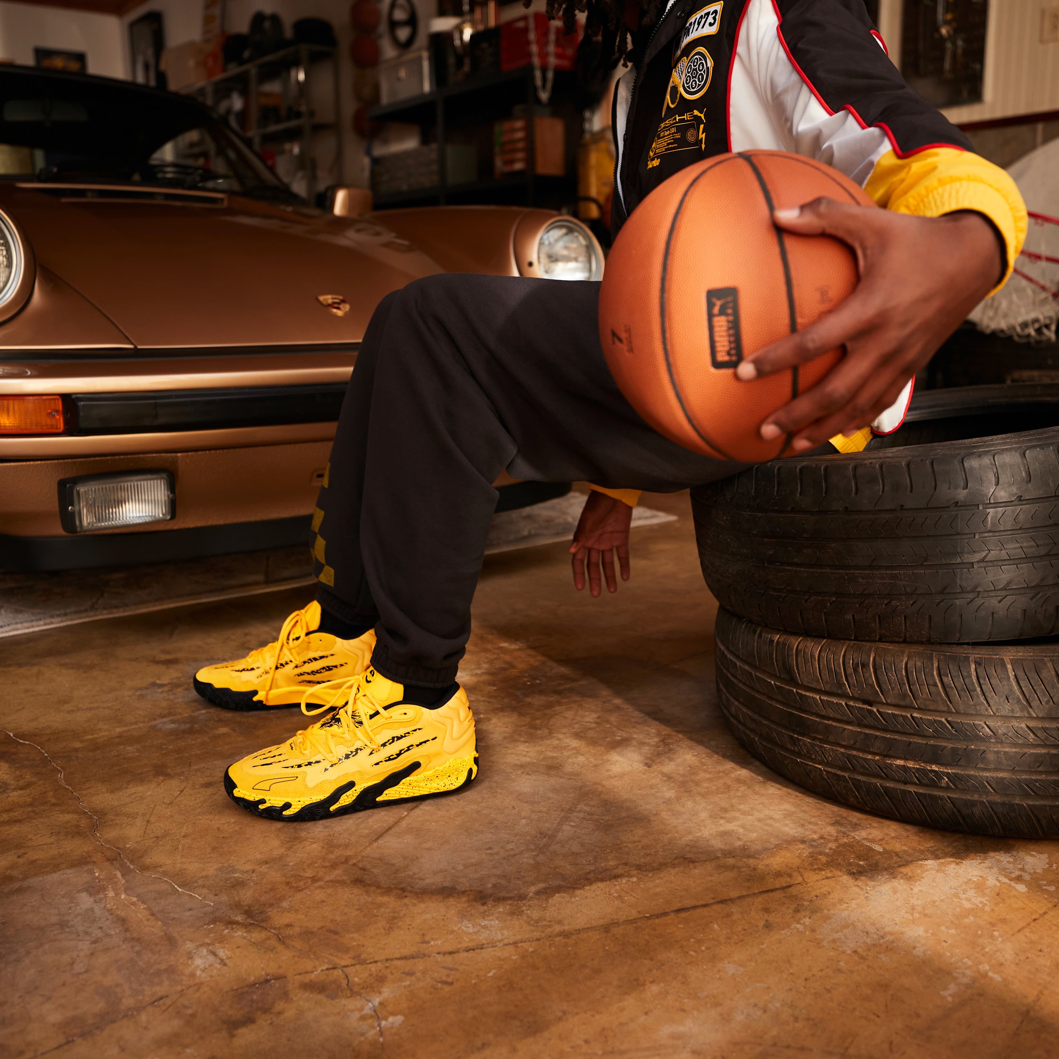 Porsche x Puma Basketball, image features man in garage dribbling basketball. He is sitting on a stack of tires with a Porsche 911 Turbo in the background.
