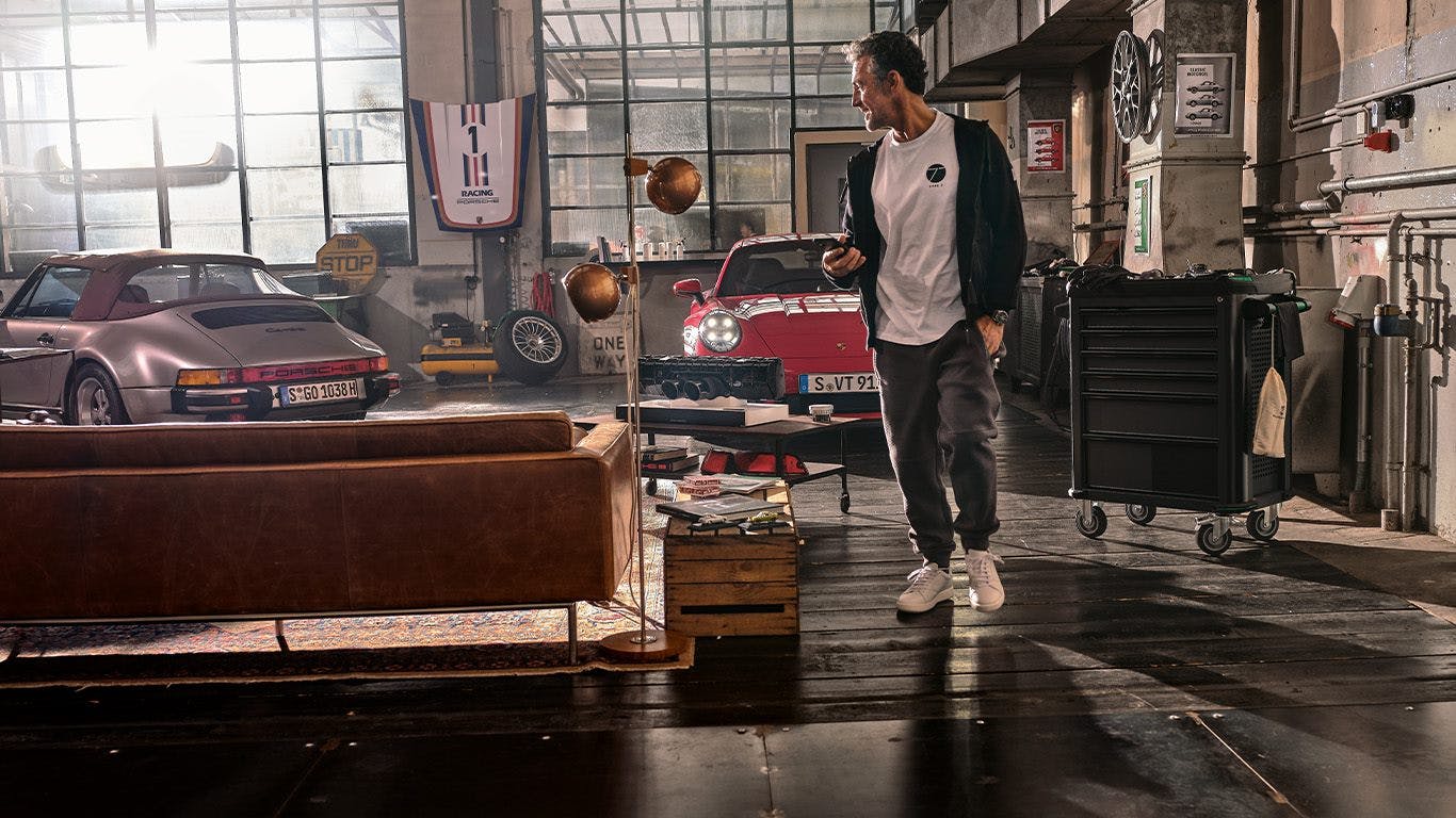 Pictured is a man in a soul garage casually looking behind the silver vintage Porsche car. Next to the classic car is a red 911. Frontally on the wall hangs a hood and laterally the rim wall clock. In the foreground of the picture is a brown leather sofa and right next to it a floor lamp.