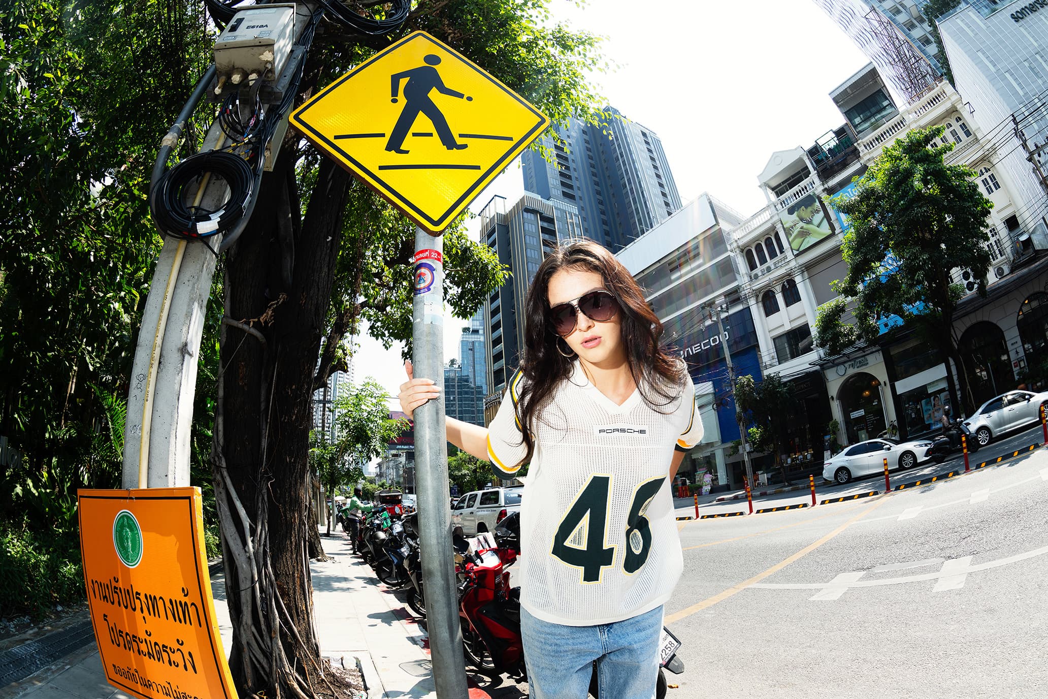 Frau mit Sonnenbrille im weißen Porsche AHEAD Football-Shirt neben einem gelben Fußgängerüberweg-Schild, Stadtstraße im Hintergrund.