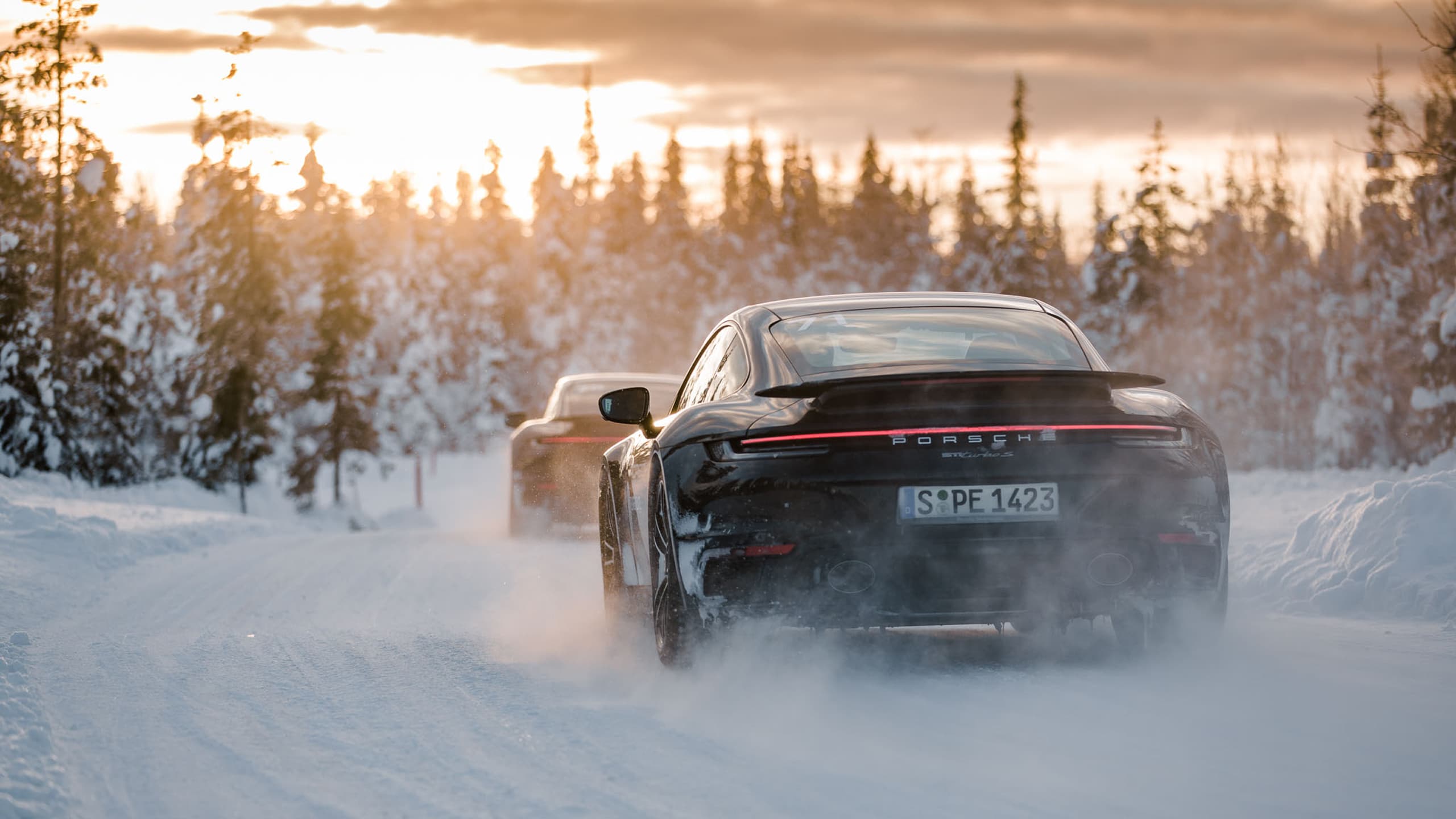 Two Porsche cars driving on a snowy road at sunset, surrounded by snow-covered trees.
