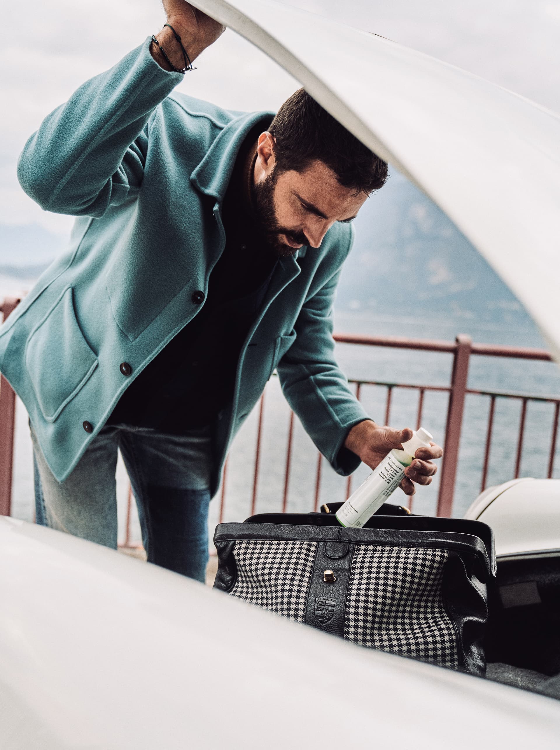 Mann in einem türkisfarbenen Mantel, der eine Flasche in eine Hahnentritt-Tasche im Kofferraum eines Autos am Seeufer legt.