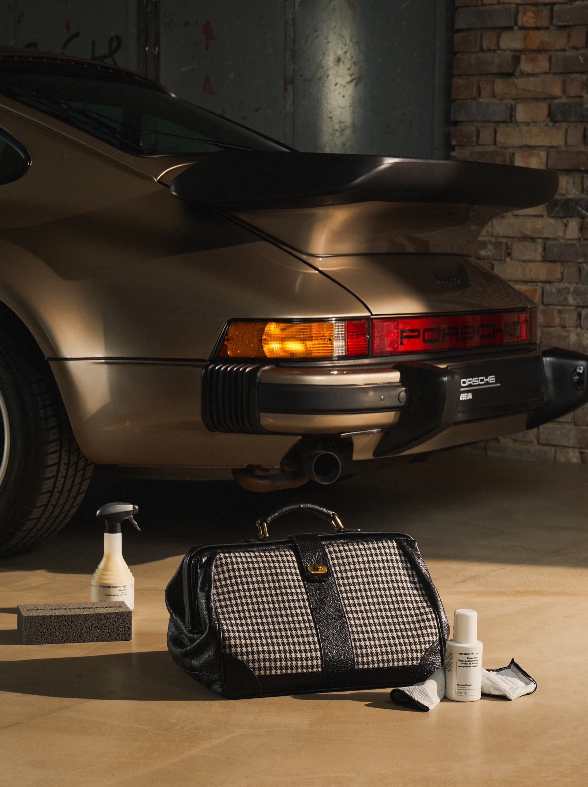 Vintage Porsche-Heck mit Hahnentritt-Tasche, Autopflegeprodukten und Reinigungstuch auf Garagenboden.