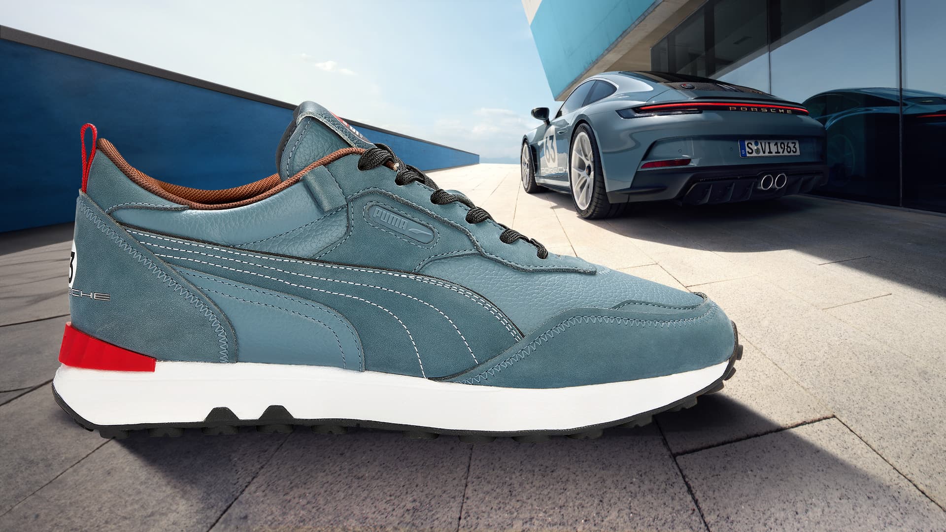 You can see a blue Puma sneaker from the side in cooperation with Porsche and a blue-coloured 911 in the background..