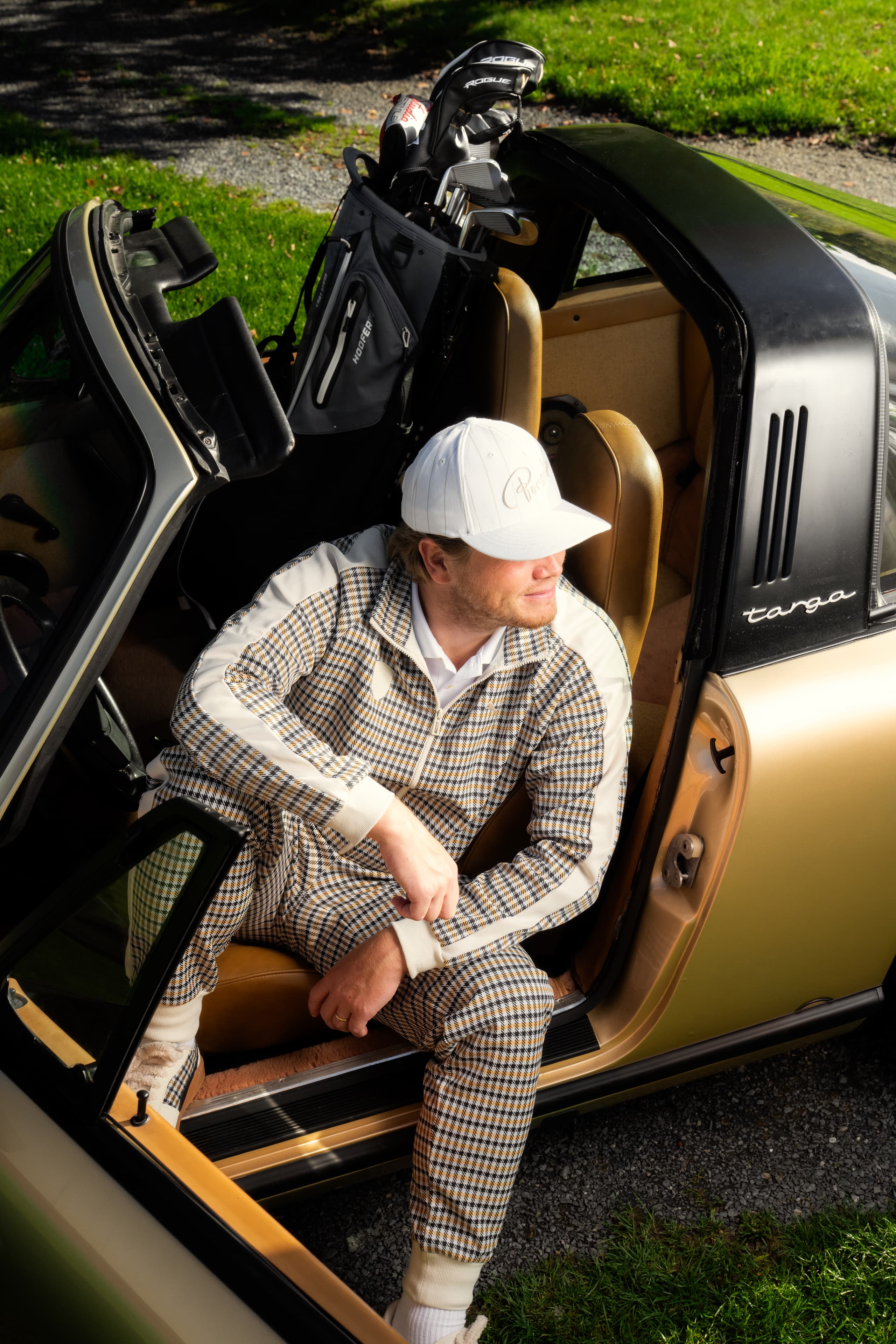 Homem com fato de treino estampado e boné branco sentado num Porsche Targa dourado descapotável, com saco de golfe atrás do banco.