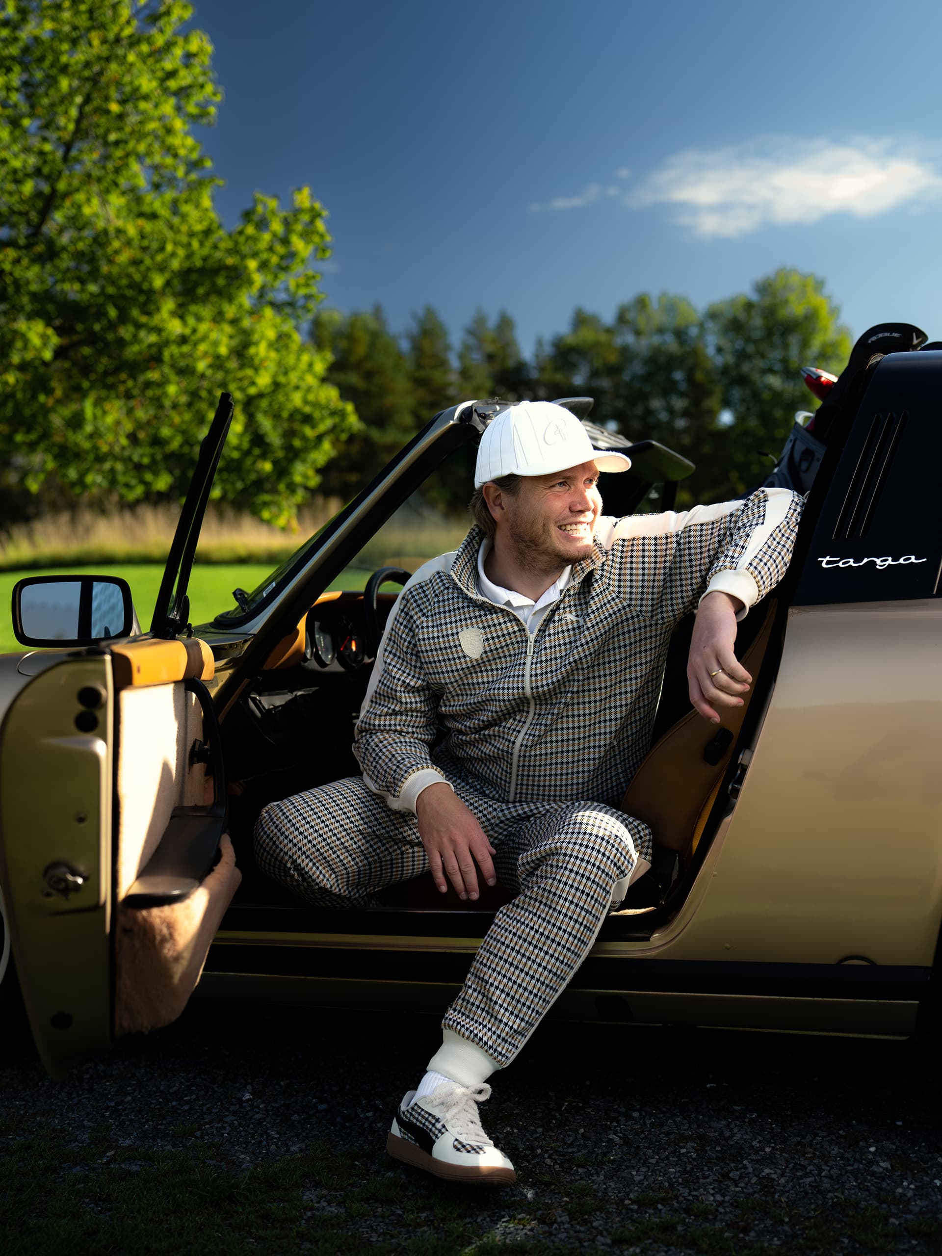 Hombre con chándal estampado y gorra blanca sentado en un Porsche Targa dorado descapotable, con una bolsa de golf detrás del asiento.