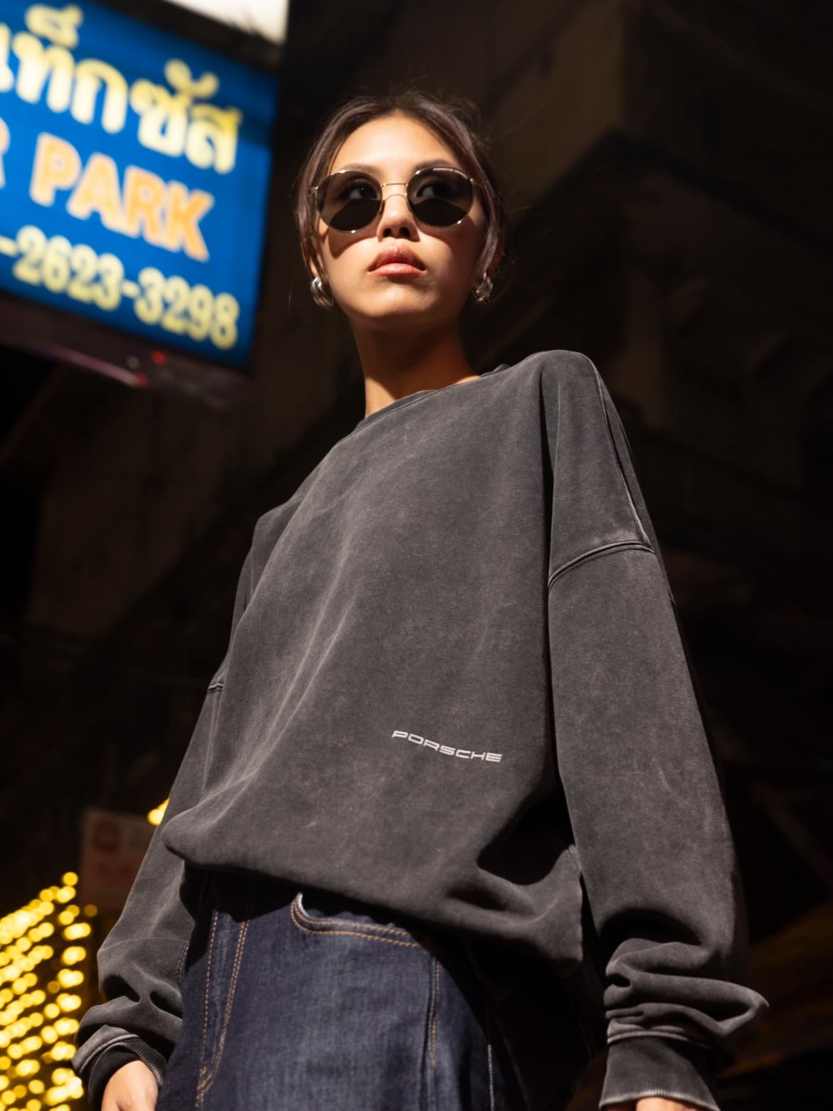 Person in dunklem Porsche-Sweatshirt und Sonnenbrille steht selbstbewusst in urbaner Nachtszene mit Neonlichtern.