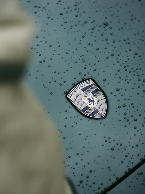 Porsche-Emblem auf einer regenbesprenkelten Motorhaube in Türkis, Nahaufnahme mit unscharfem Vordergrund.