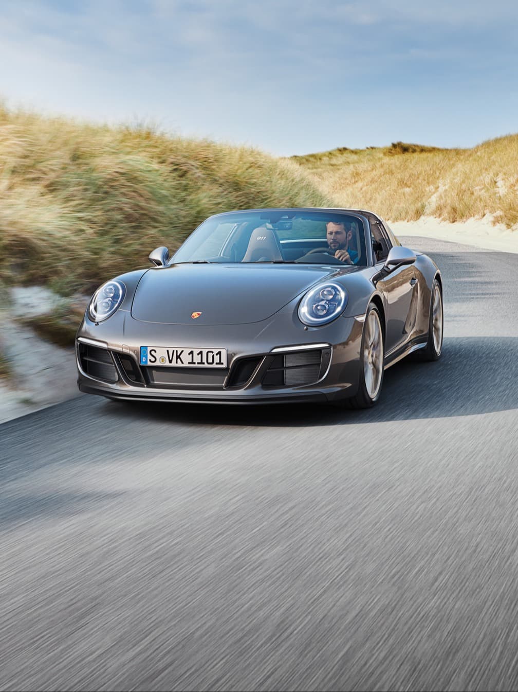 Cabriolet Porsche 911 cinzento a circular numa estrada costeira entre dunas, condutor ao volante, céu azul.