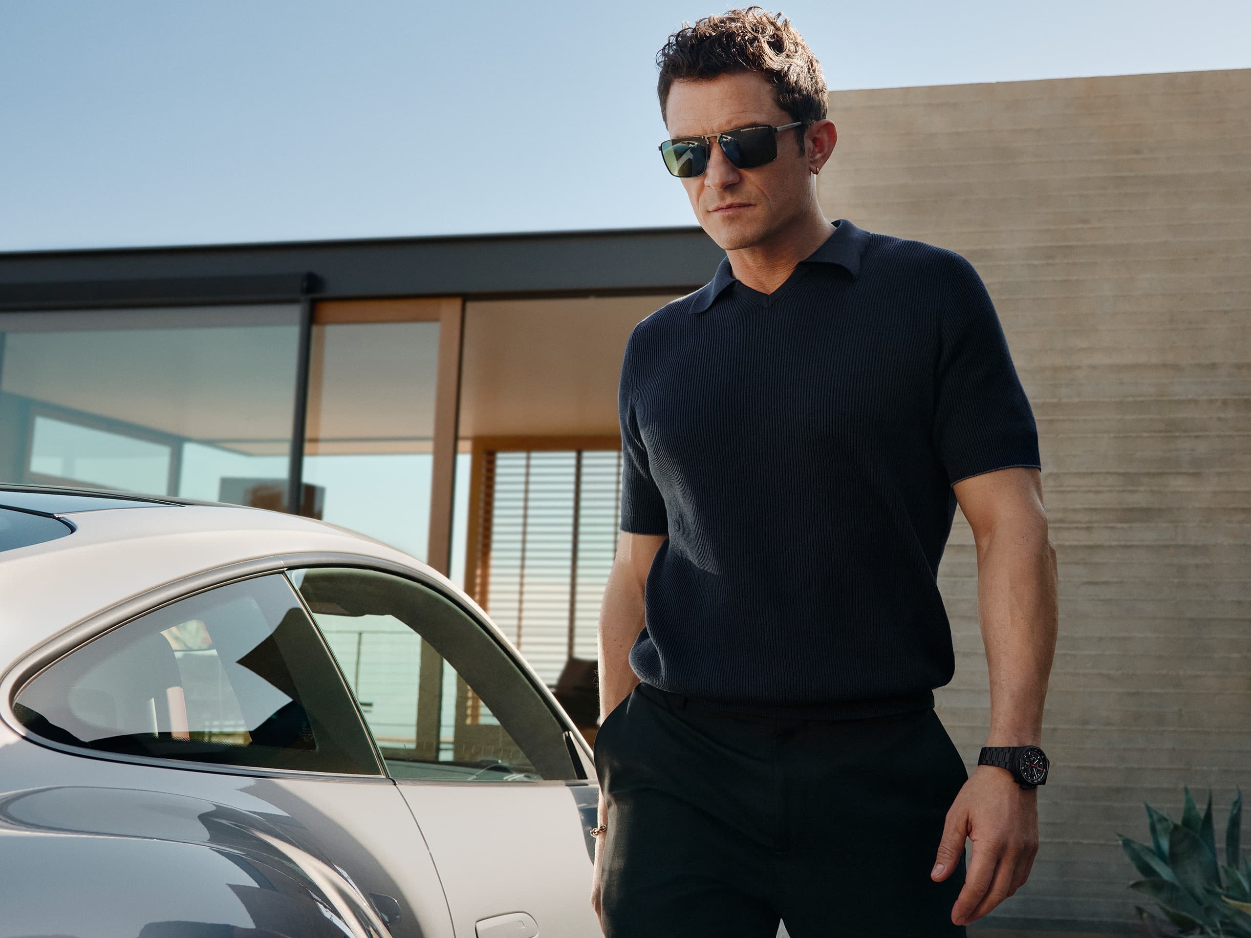 Orlando Bloom ma na nadgarstku zegarek Chronograph 1 – All Black Numbered Edition, idzie w zadaszonym miejscu parkingowym, obok stoi jego Porsche