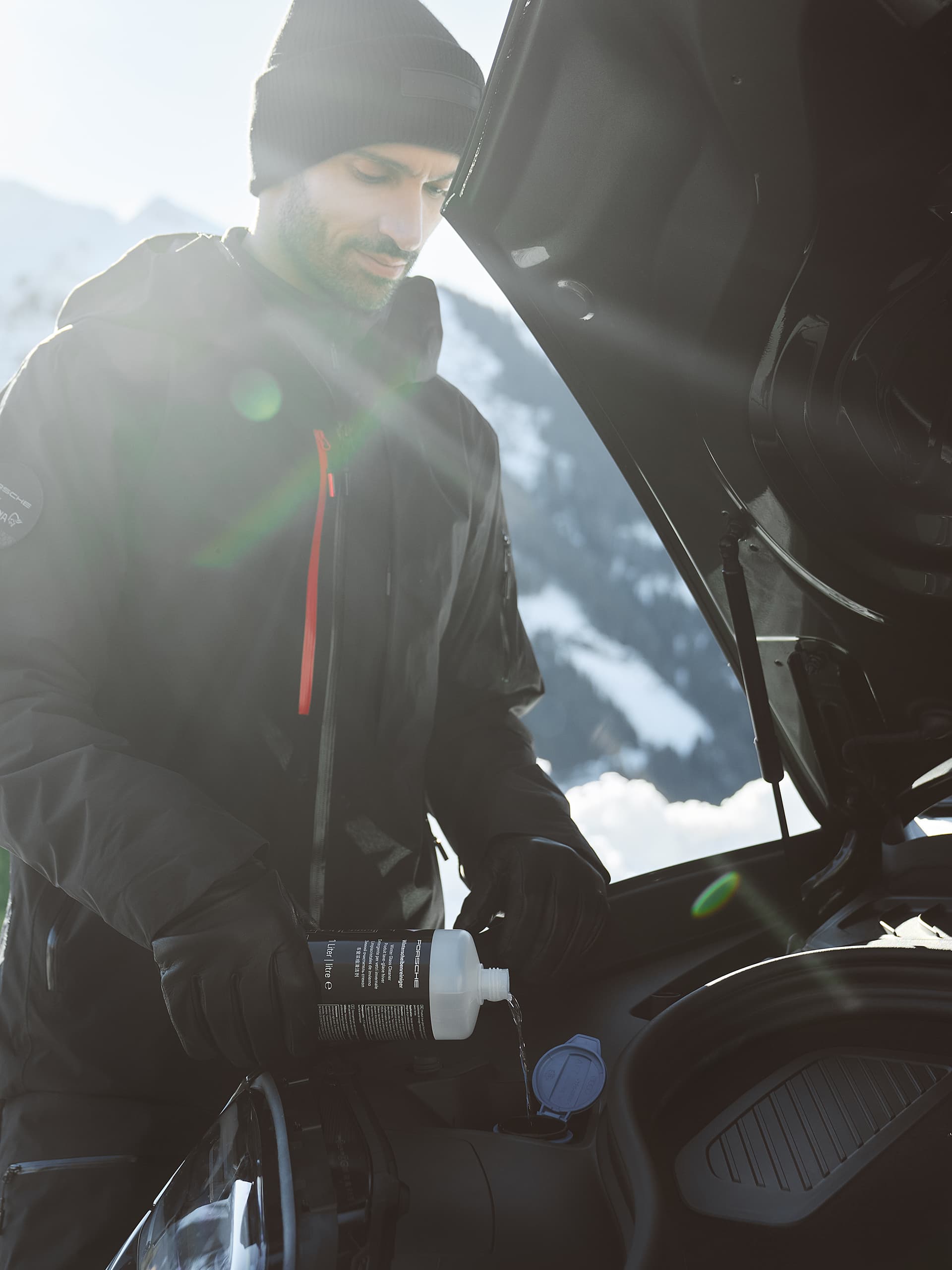 Mann in schwarzer Jacke und Mütze gießt Flüssigkeit in den Motor eines Autos, schneebedeckte Berge im Hintergrund, Sonnenlicht strömt.