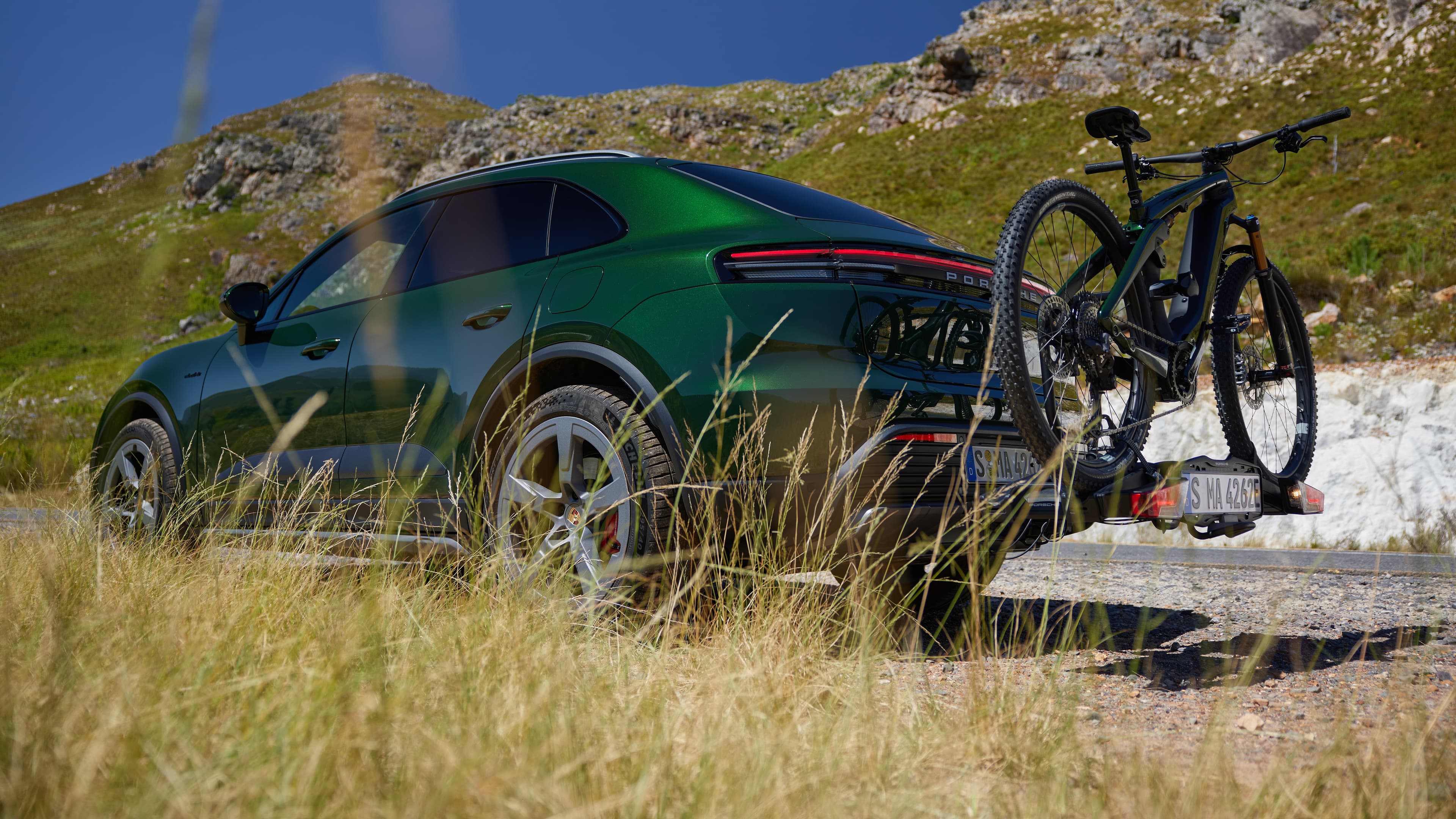 Green Porsche SUV with a bike rack on a mountain road, surrounded by grass and rocky hills under a clear blue sky.