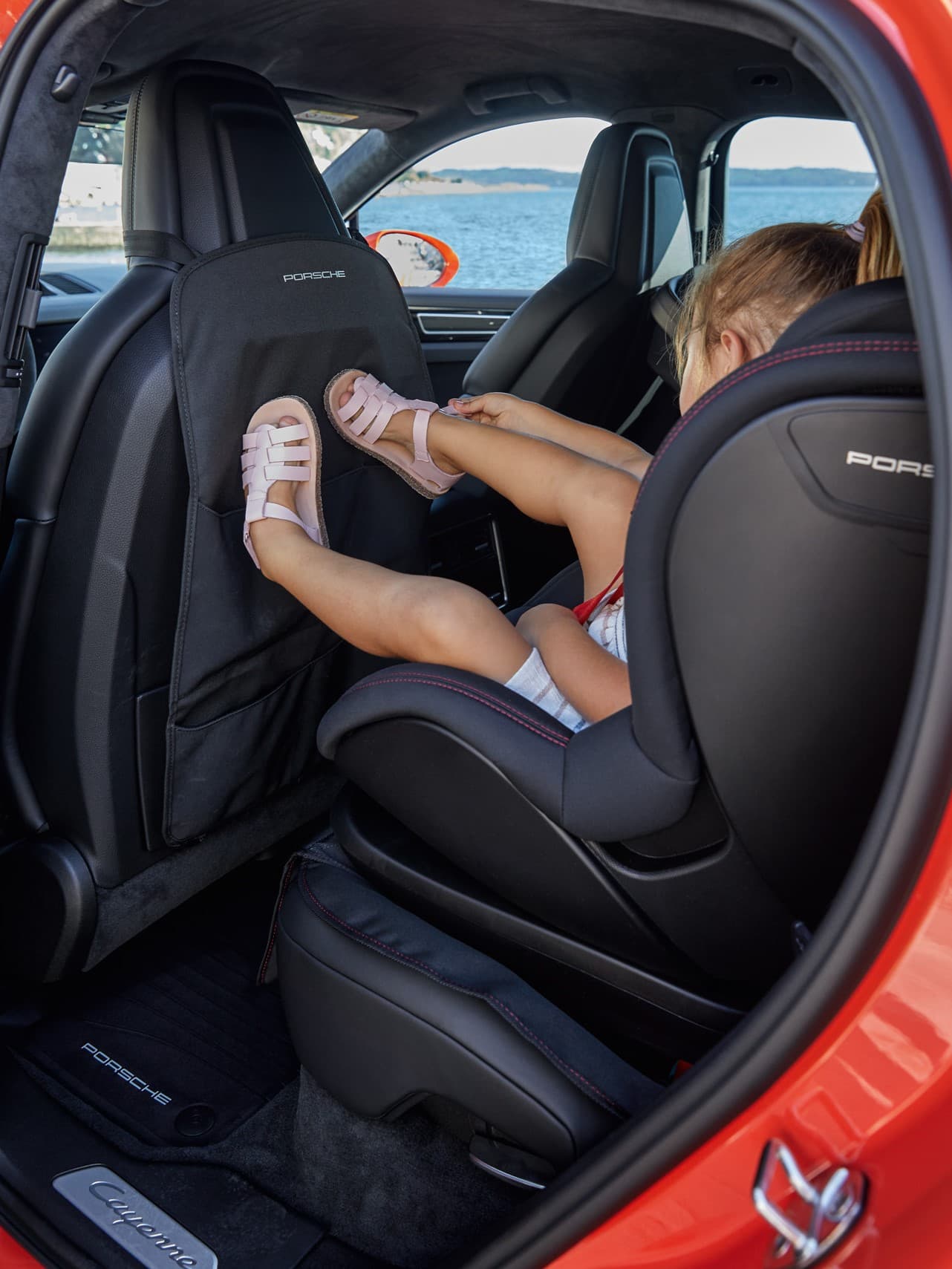 Enfant dans un siège auto donne des coups au dos du siège avant dans une Porsche rouge, vue sur le lac à l'extérieur.