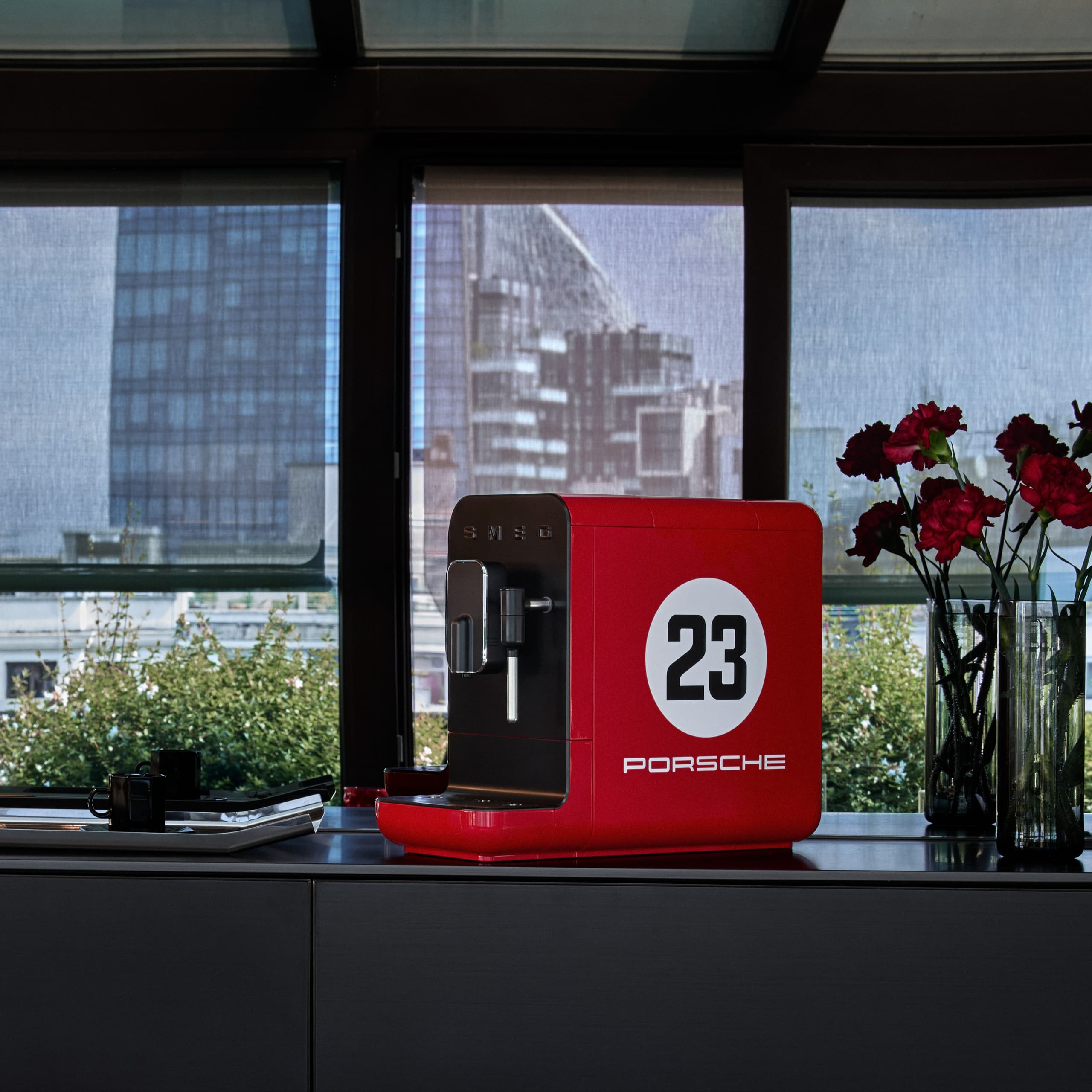 Machine à café Porsche rouge avec le numéro 23, sur un comptoir à côté de fleurs rouges, avec une vue urbaine en arrière-plan.
