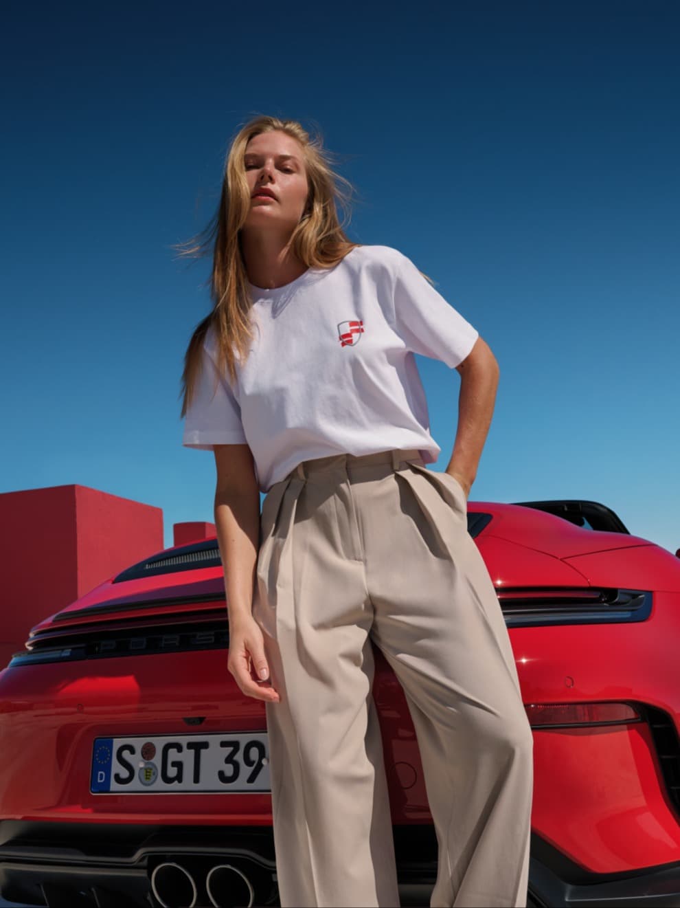 Woman in a white T-shirt and beige trousers standing in front of a red Porsche convertible, blue sky in the background.