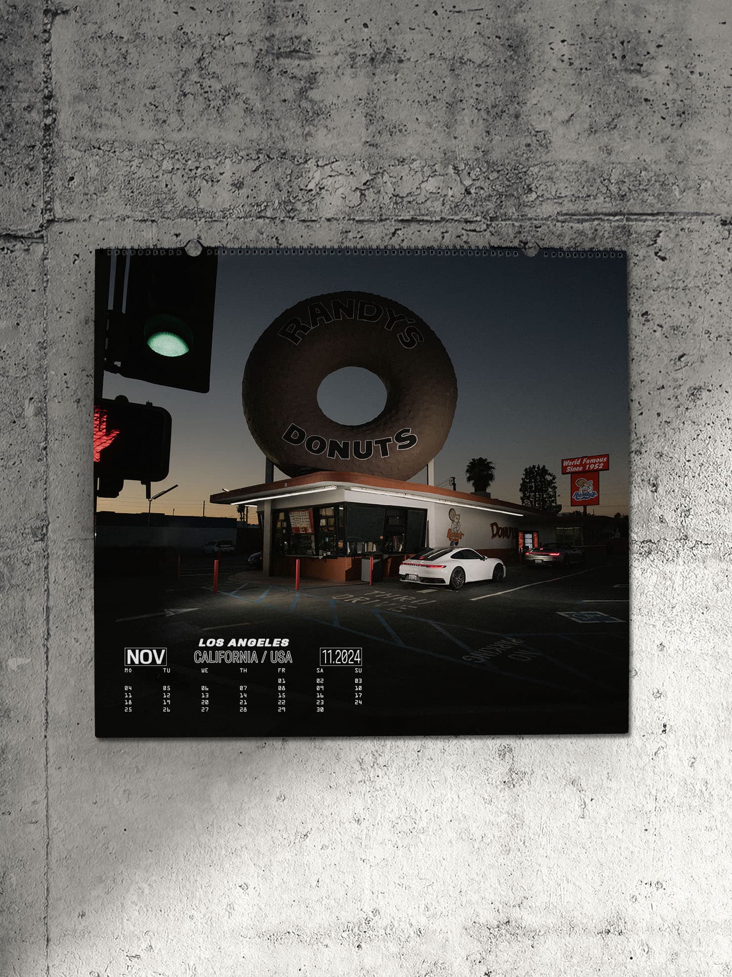 Pictured is a Porsche calendar on a gray concrete wall. It shows a night situation with a supermarket, which has a large donout on the roof. Parked next to it is a white Porsche 911.