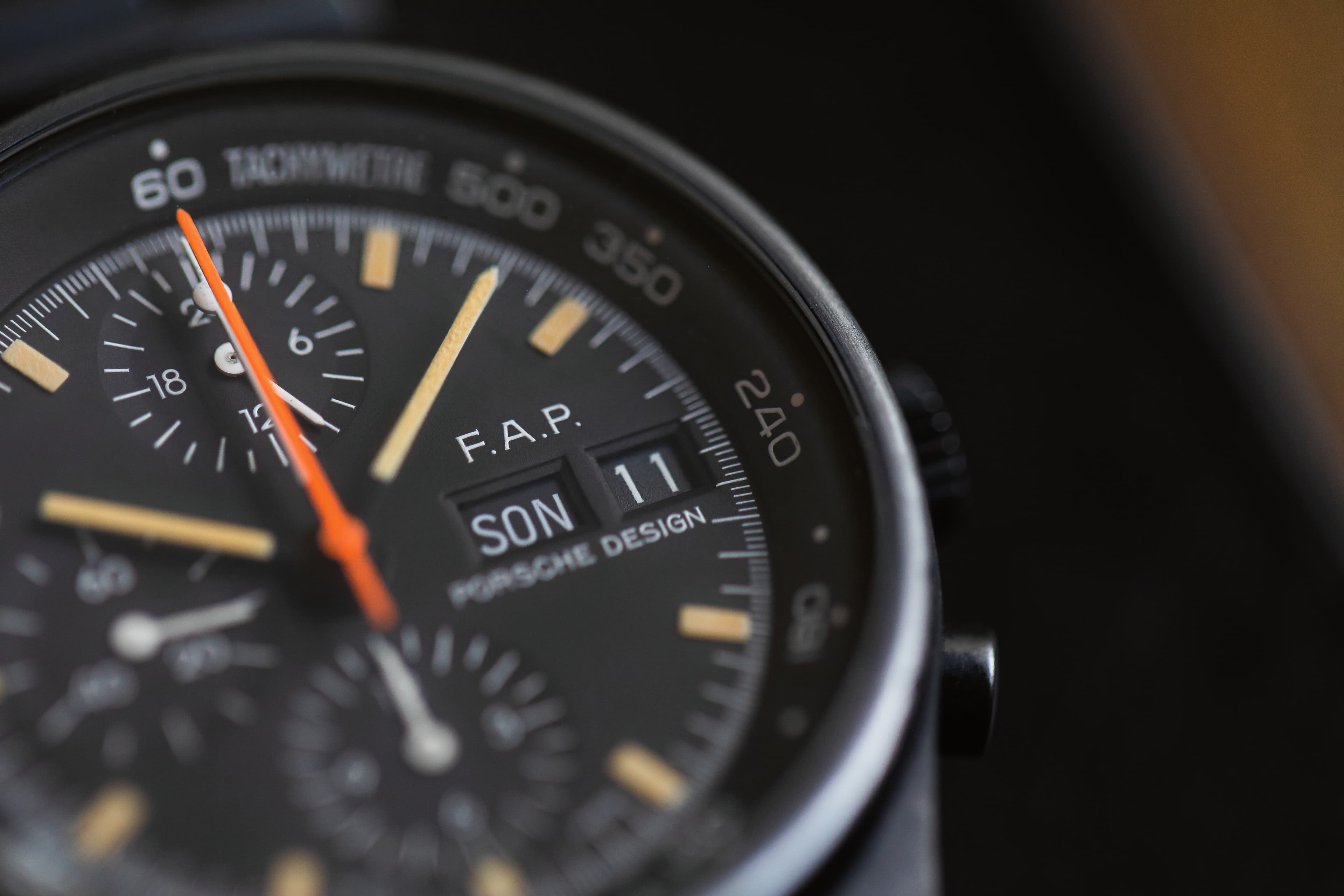 Close-up of a black Porsche Design chronograph with orange second hand and date display.
