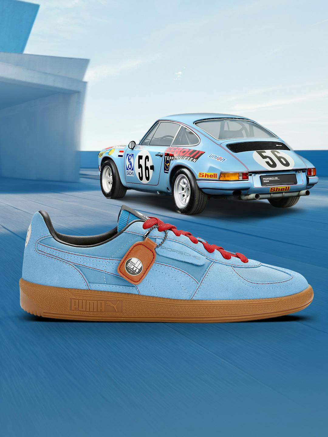 You can see a flat blue Puma sneaker from the side with a brown rubber sole in cooperation with Porsche and in the background there is a blue-coloured vintage car with colourful decorations.