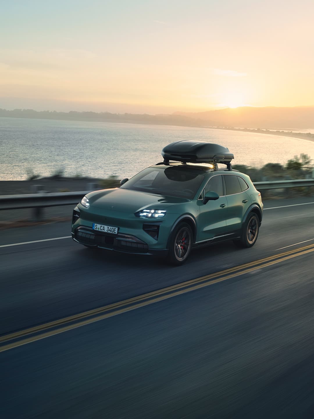 Green Porsche SUV with a black roof box driving along a coastal road at sunset.