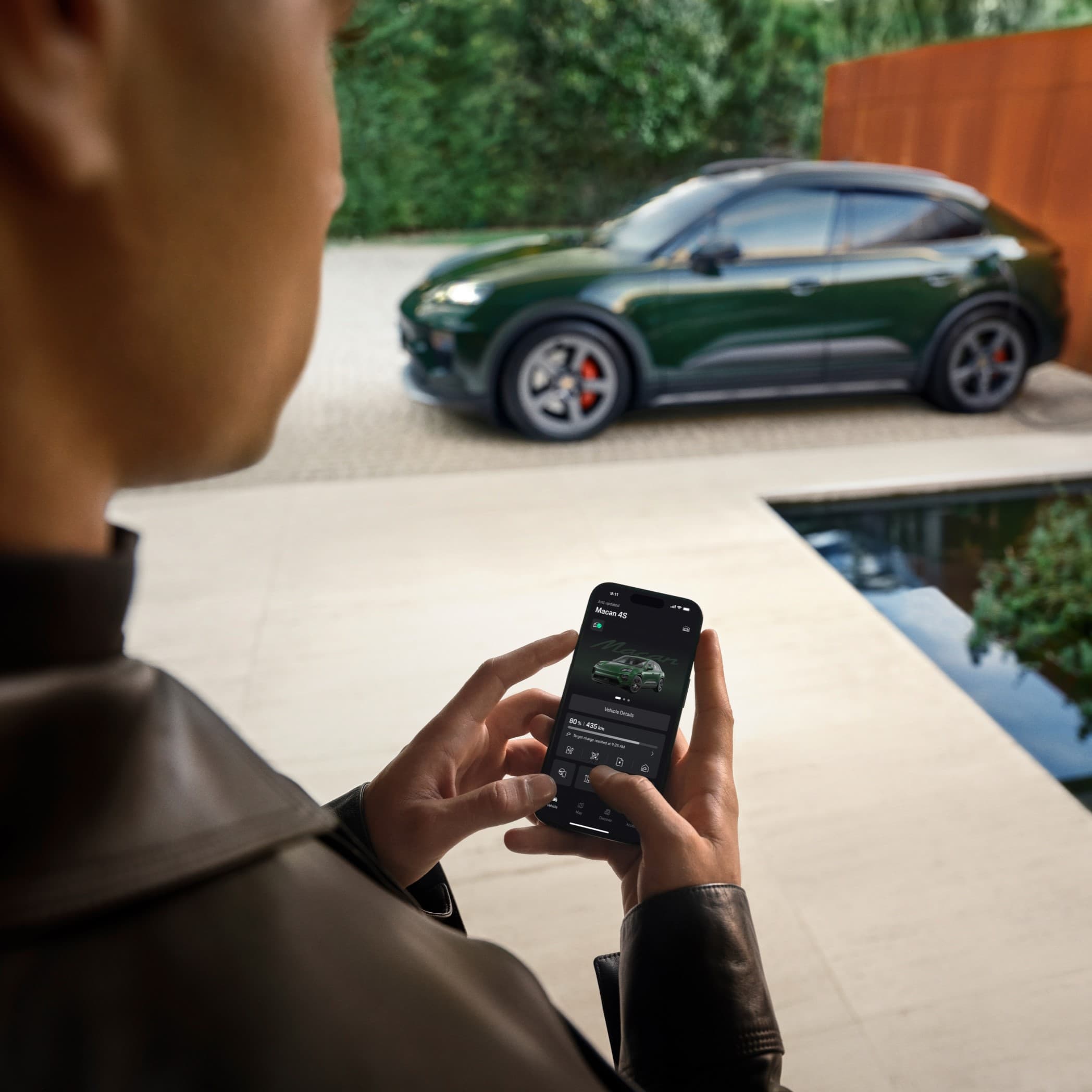 Person using smartphone app to control green Porsche Macan parked outside, with modern architecture and greenery in background.