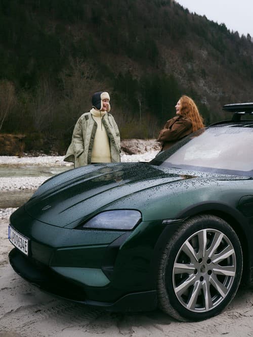 Grüner Porsche am Fluss geparkt, zwei Personen in Winterkleidung stehen in der Nähe, bewaldete Hügel im Hintergrund.