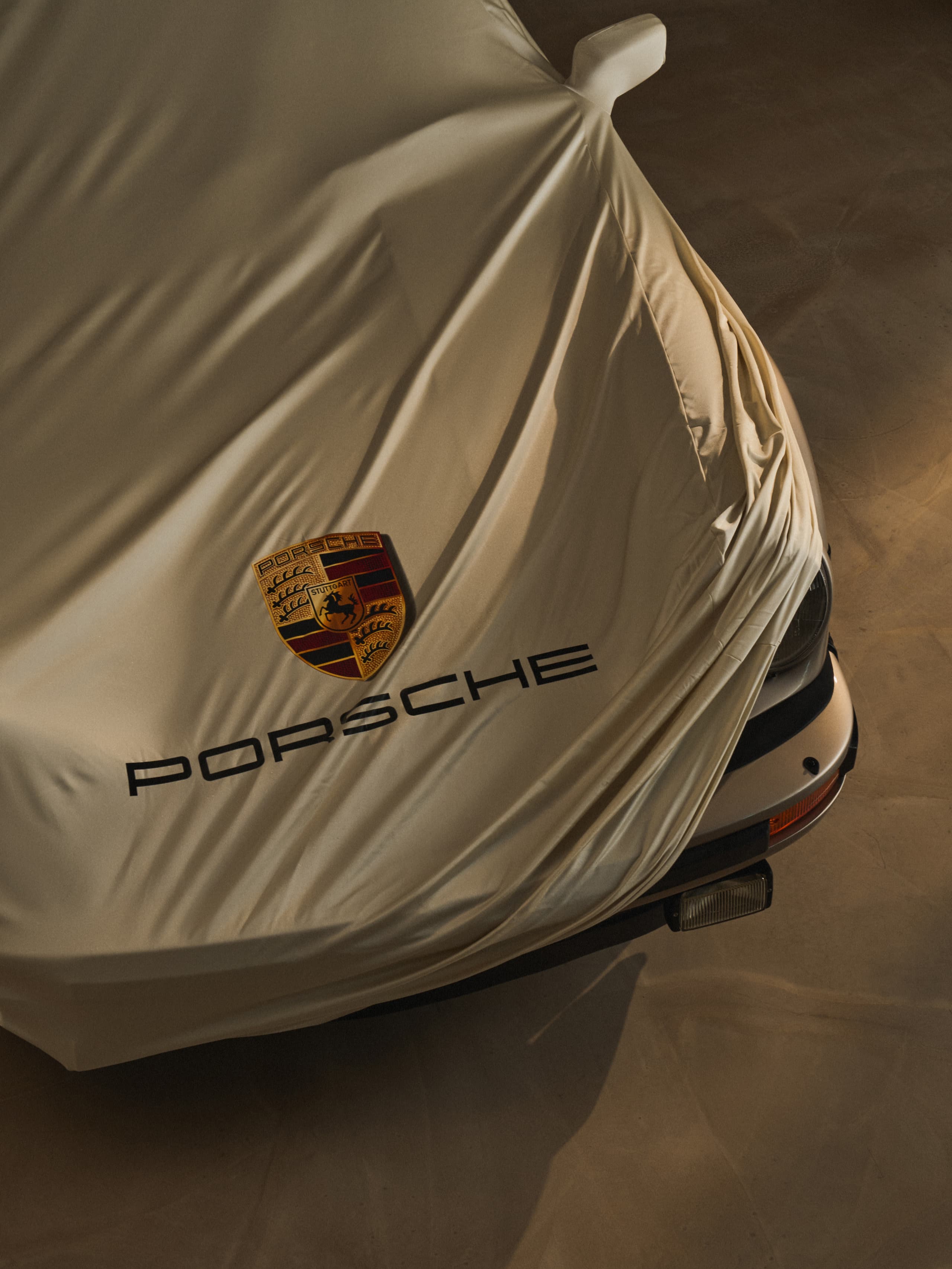 Car covered with beige fabric, displaying Porsche logo and text, on a dimly lit surface.