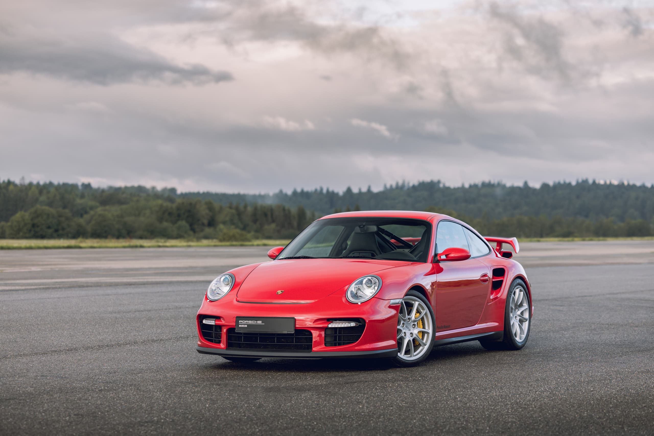 Porsche 911 rouge sur une surface asphaltée, avec grand aileron arrière, devant une forêt et sous un ciel nuageux.