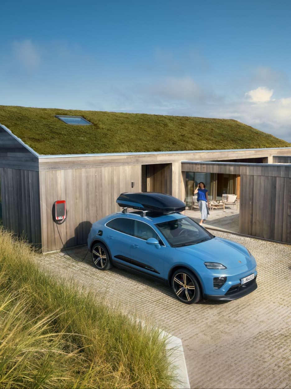 Blue Porsche SUV with black roof box charging at a wallbox in front of a wooden house with a green roof; person on the terrace.