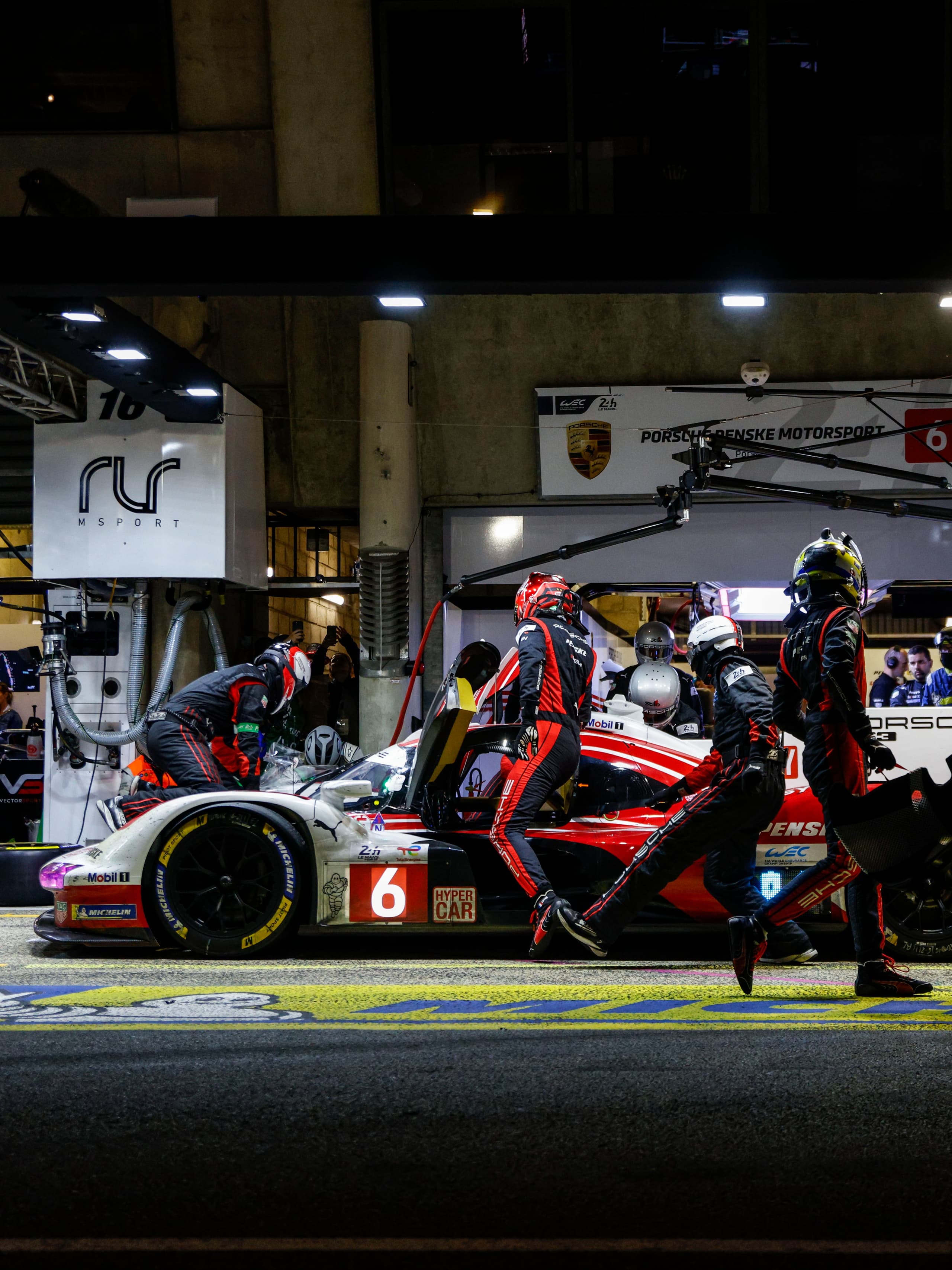 Produktdetaljsider: Pitcrew i svarte og røde dresser utfører service på en rød og hvit Porsche-racerbil under et nattlig depotstopp