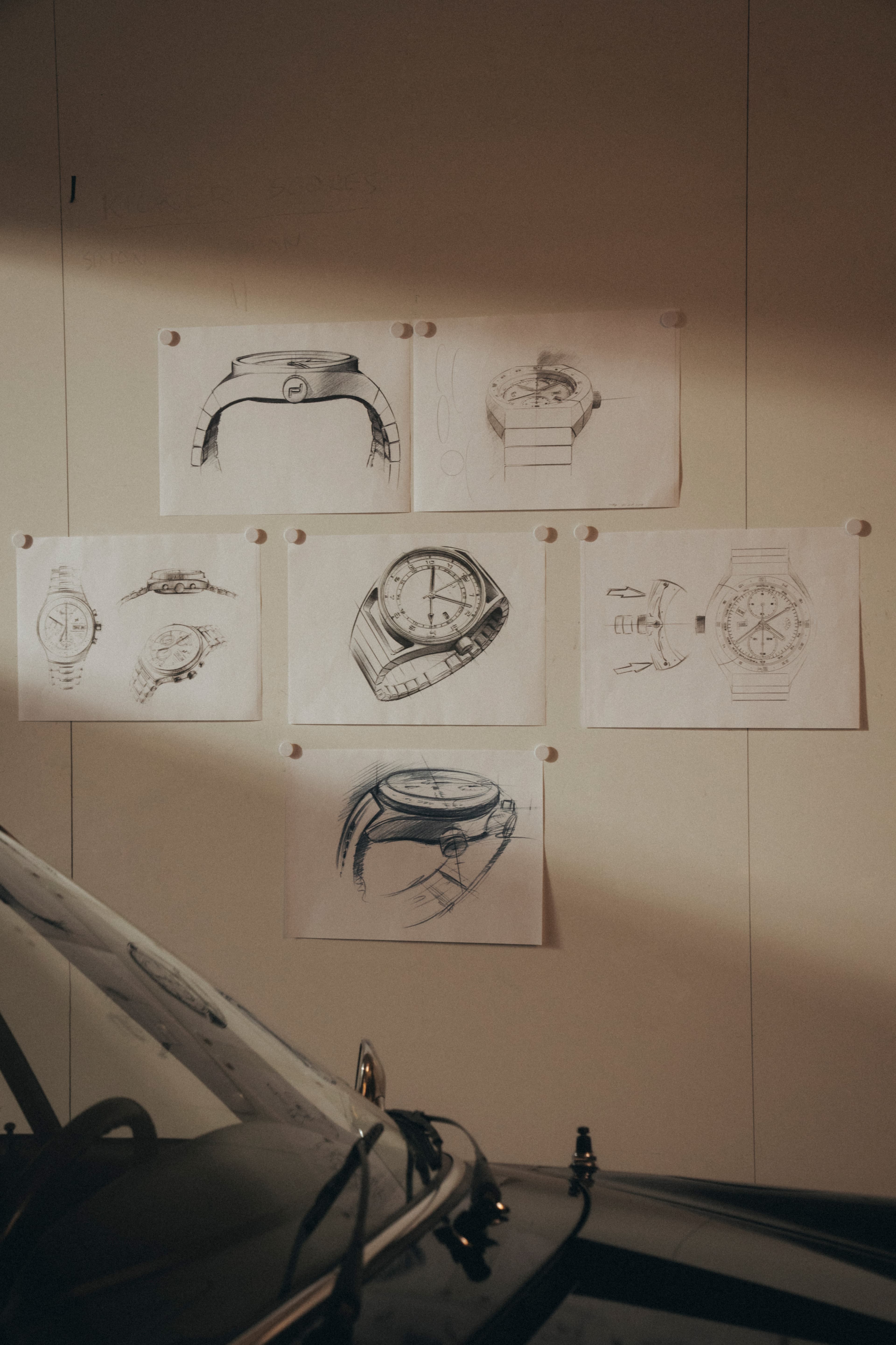 Skizzen von Armbanduhren auf Papier, mit Pins an einer Wand befestigt; unten unscharfer Autoteil im Vordergrund.