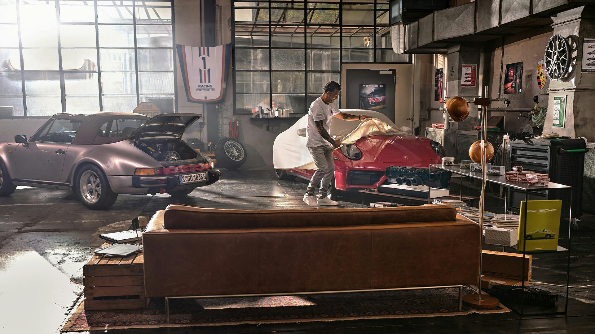 Pictured is a Soul Garage with a man covering a red 911. On the left of the picture is a vintage Porsche car. In the foreground is a brown leather sofa and to the right is a floor lamp. Frontally on the wall there is a hood and on the right side of the wall there is a rim wall clock. Soul Garage is equipped with many accessories like a soundbar or a sideboard with model cars.