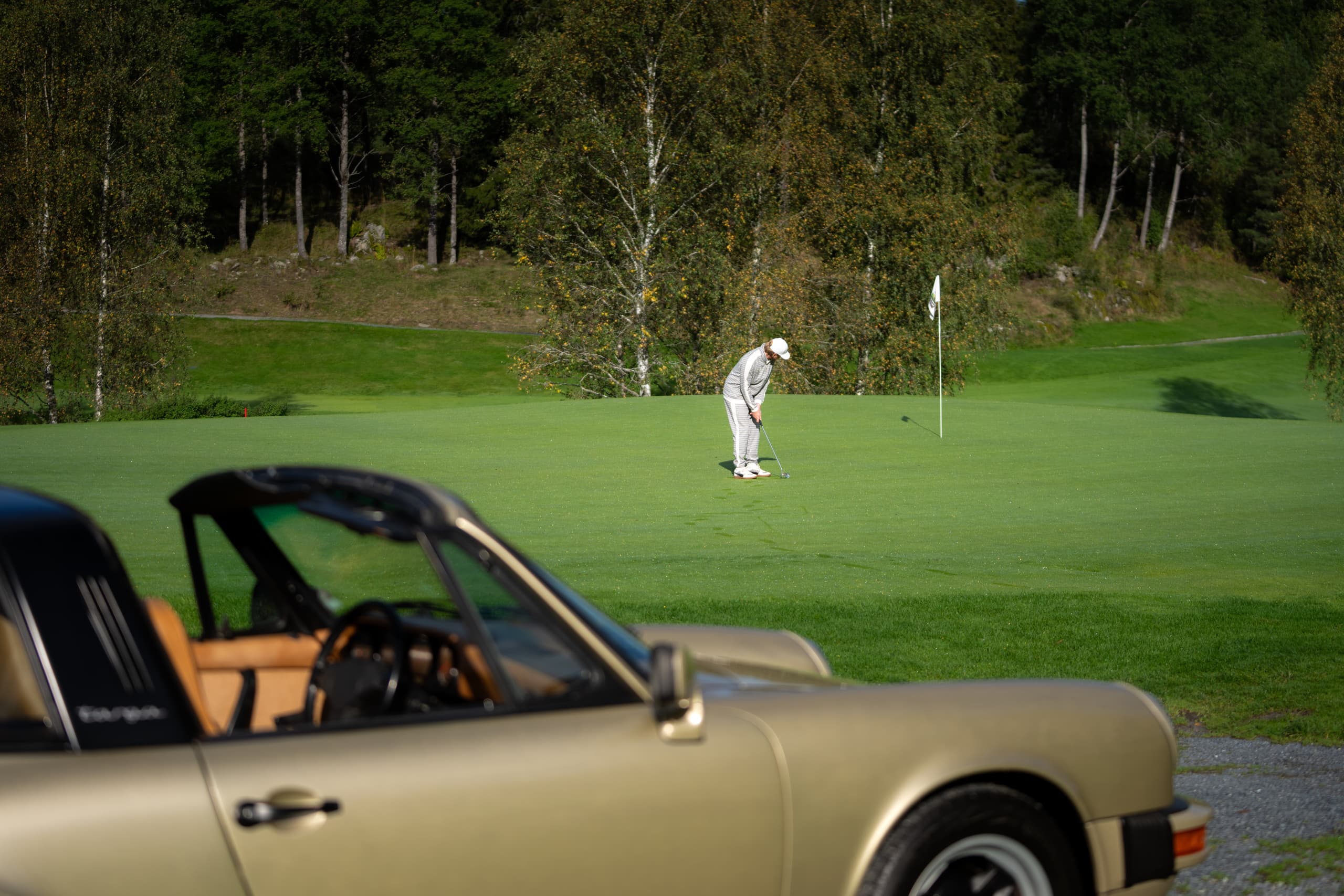 Golfer in Weiß puttet auf einem grünen Platz, mit einem goldenen Vintage-Porsche im Vordergrund, umgeben von Bäumen.
