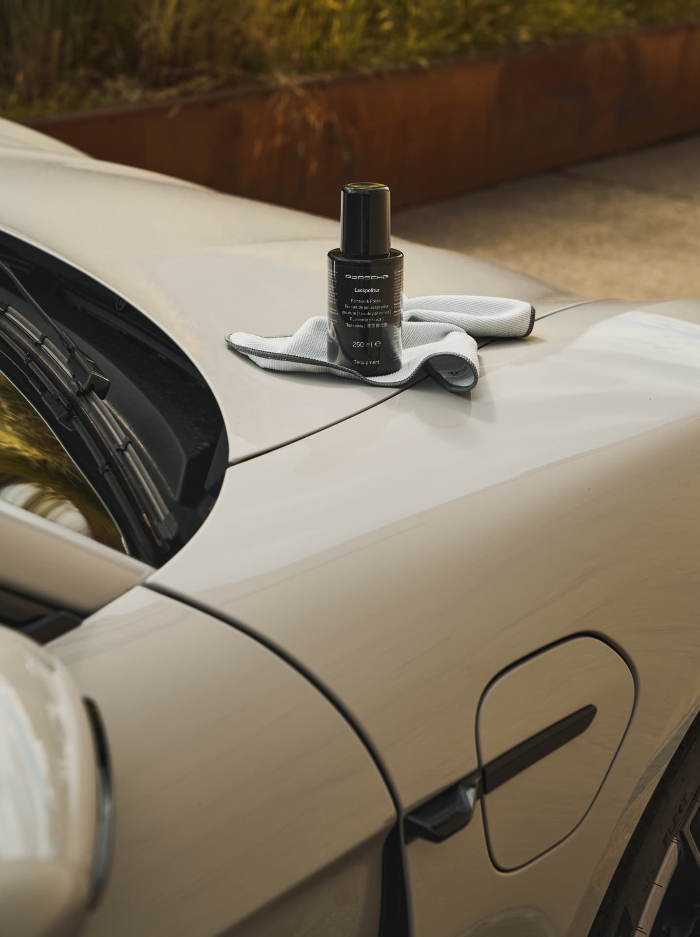 Porsche polish bottle on a gray car hood with a microfiber cloth, outdoors with blurred greenery in the background.
