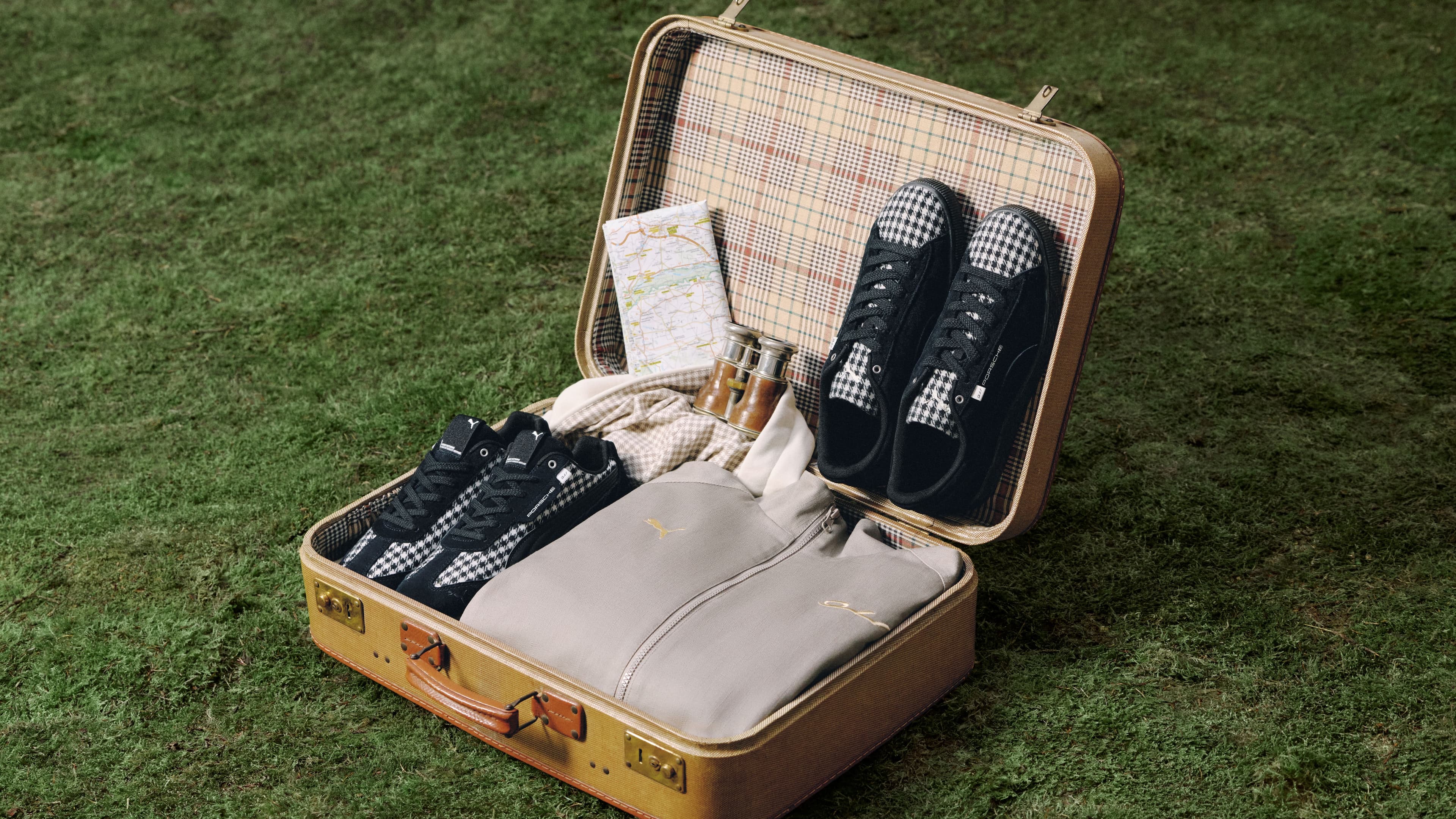 Open beige suitcase on grass with black and white checkered sneakers, map, binoculars, and a gray bag