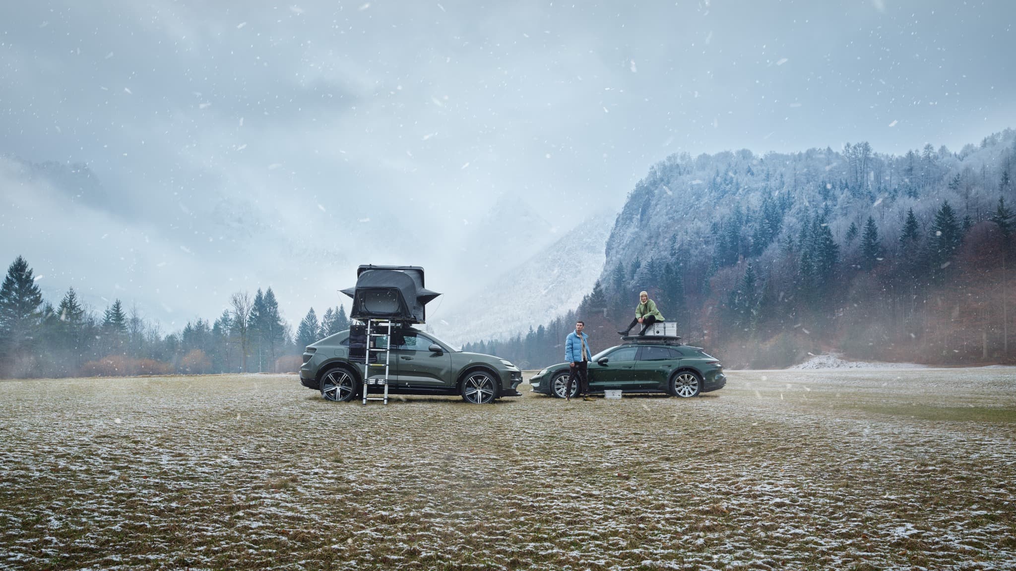 Two SUVs with roof tents in snowy landscape, people standing and sitting, surrounded by snow-dusted trees and mountains.