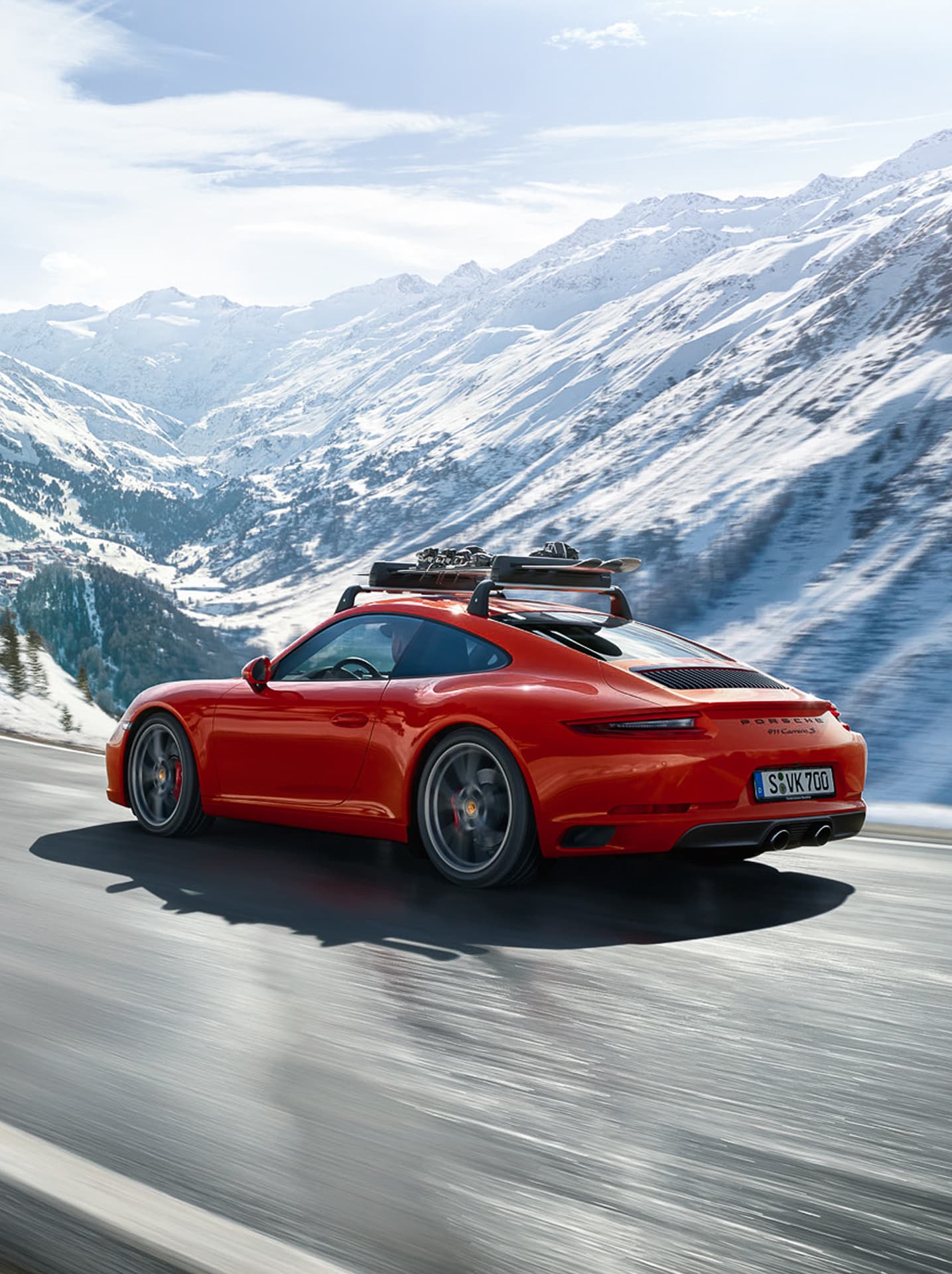 Red Porsche 911 Carrera with roof rack driving on snowy mountain road under clear sky.