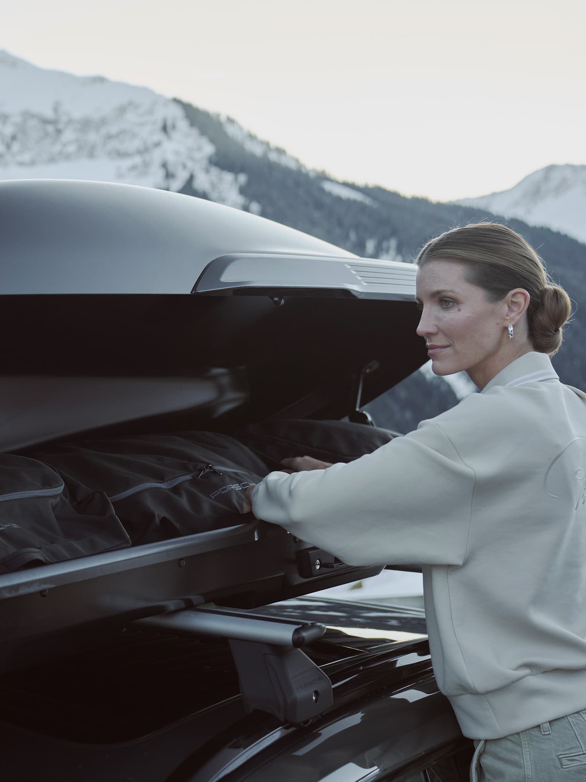 Woman loading black bag into car roof box, snowy mountains in background.