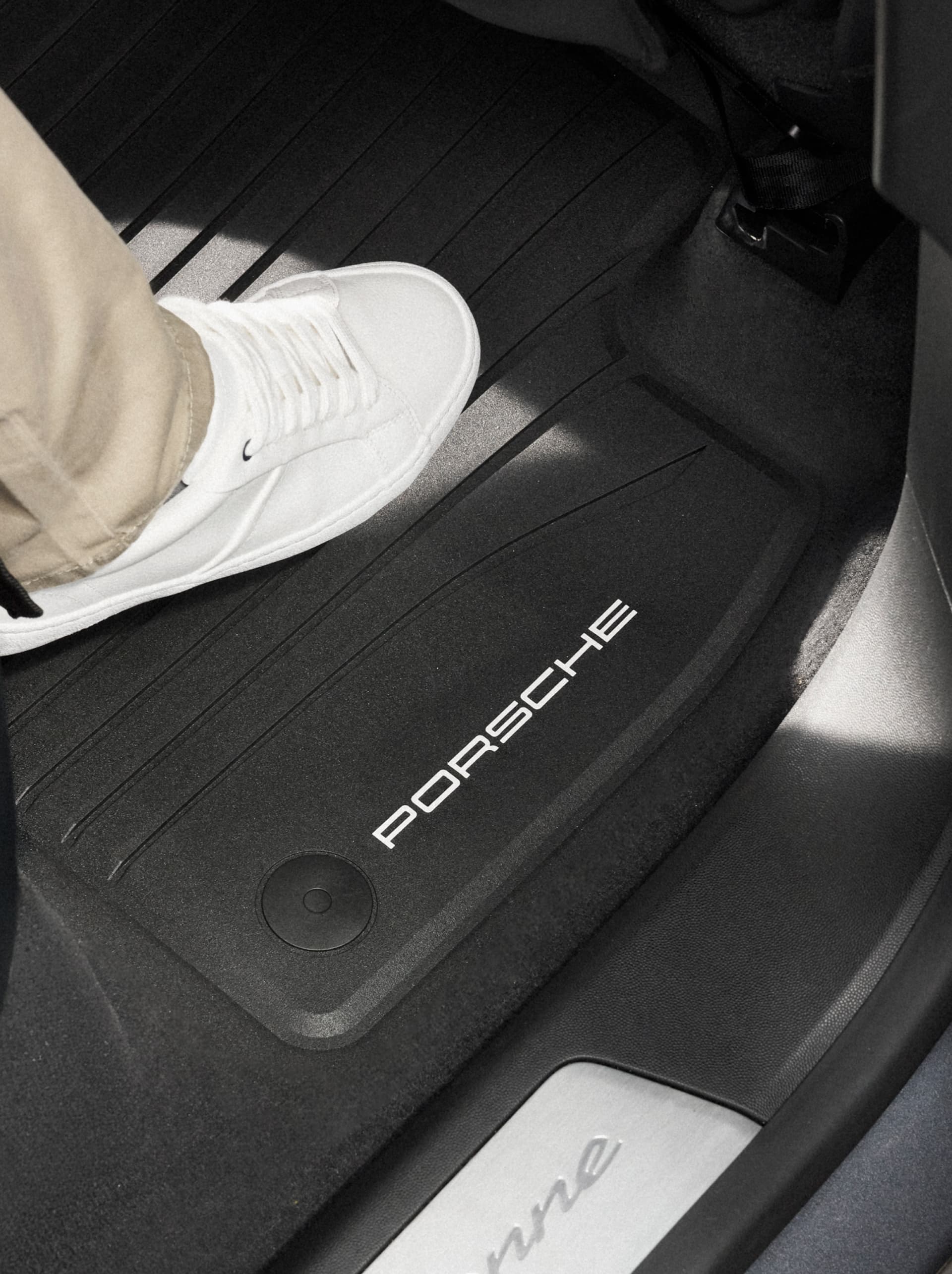 White sneaker stepping on black Porsche all-weather floor mat with PORSCHE logo in car footwell