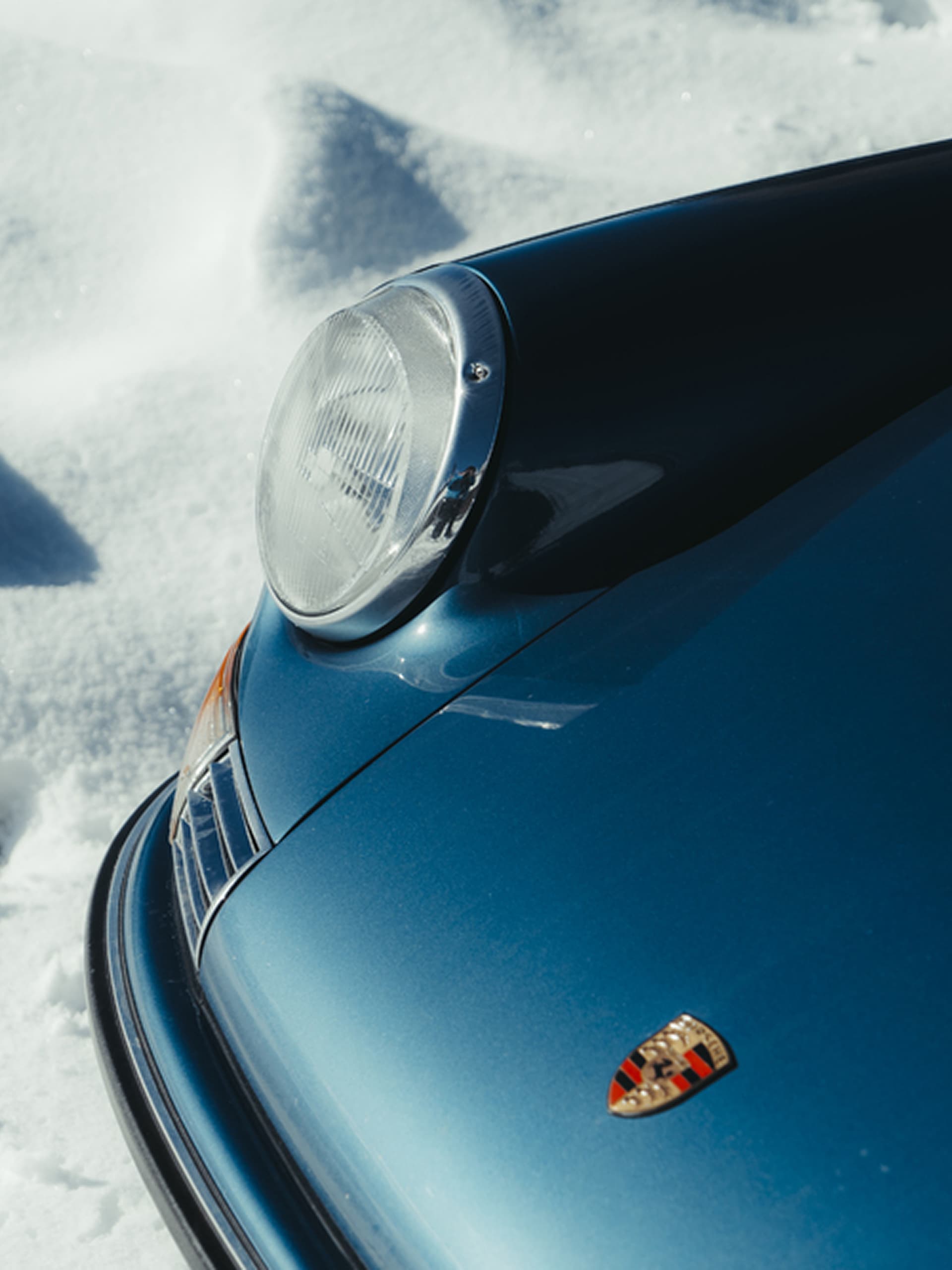 Blauer Porsche mit Frontscheinwerfer und Emblem, auf Schnee geparkt.