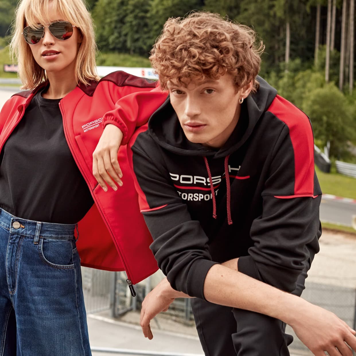 Twee modellen in Porsche Motorsport-kleding, rode jas en zwarte hoodie, buiten met een racebaan op de achtergrond.
