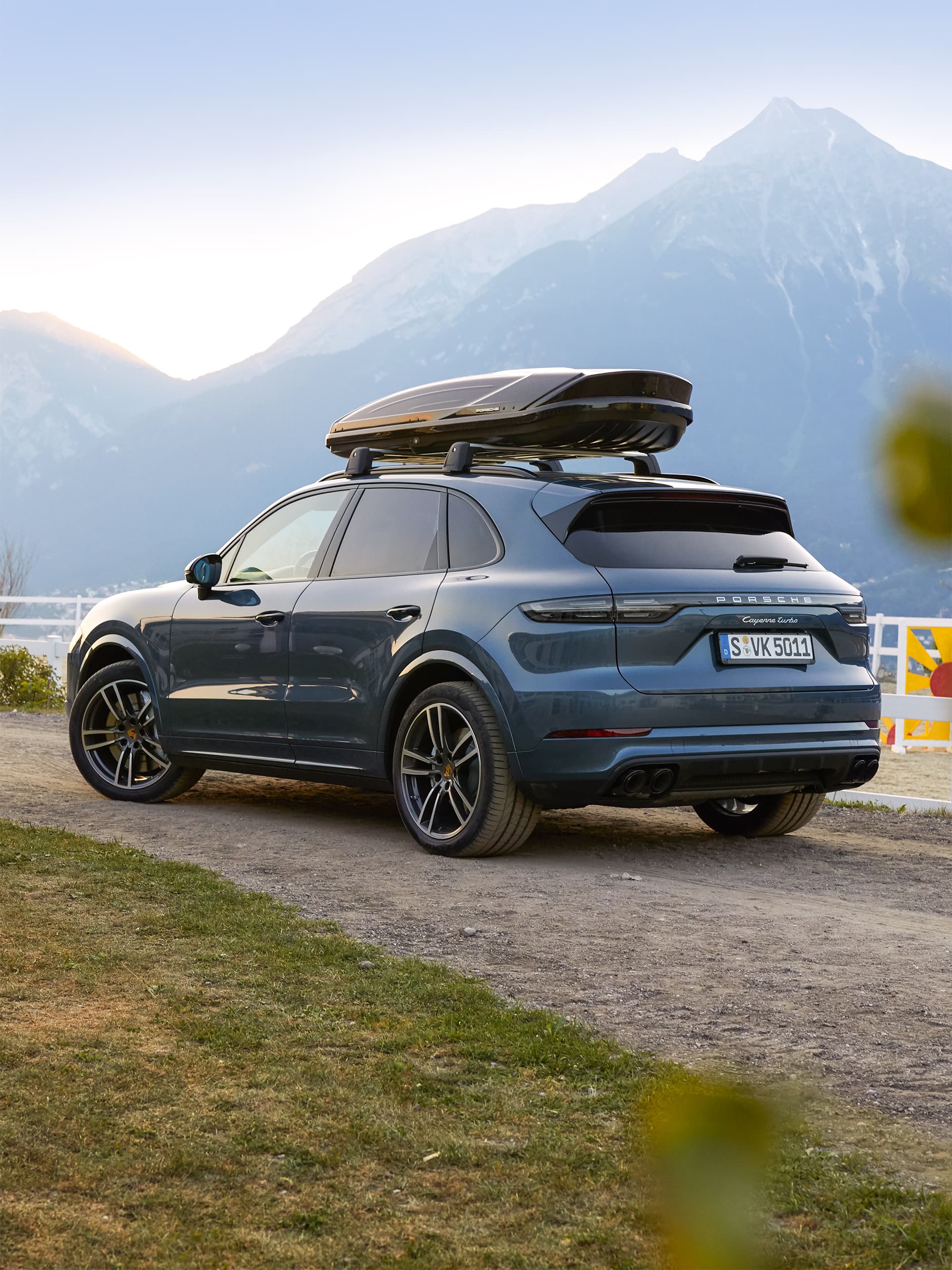 Porsche Cayenne gris avec coffre de toit noir, stationné sur un chemin de gravier devant un panorama de montagnes à la lumière du soir.