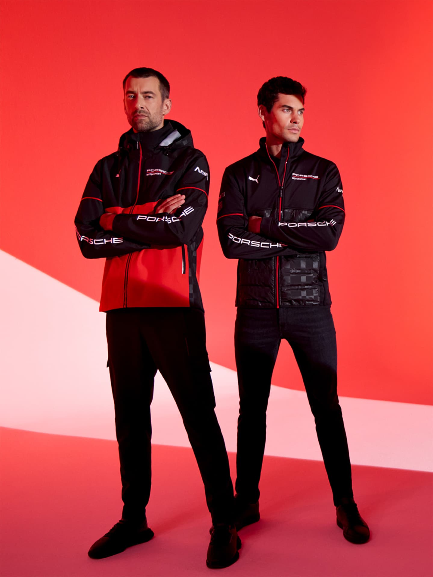 Two men in black and red Porsche jackets stand confidently against a vibrant red background.