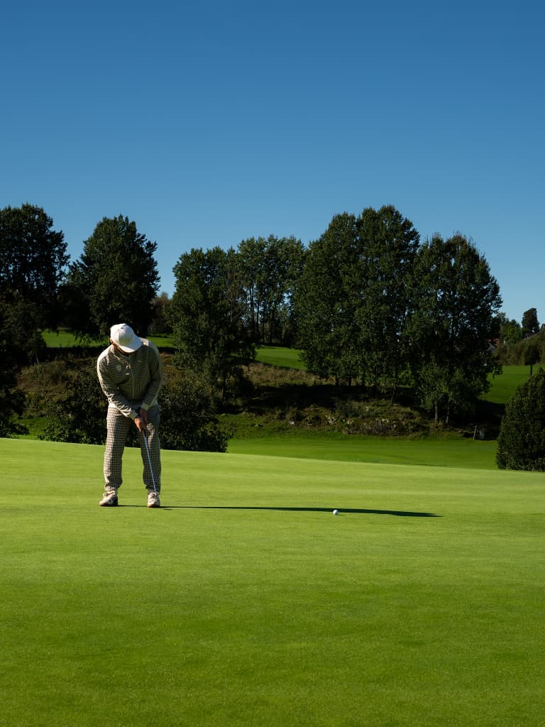 Golfer in beige outfit and white cap putting on a green fairway under clear blue sky, trees in the background.