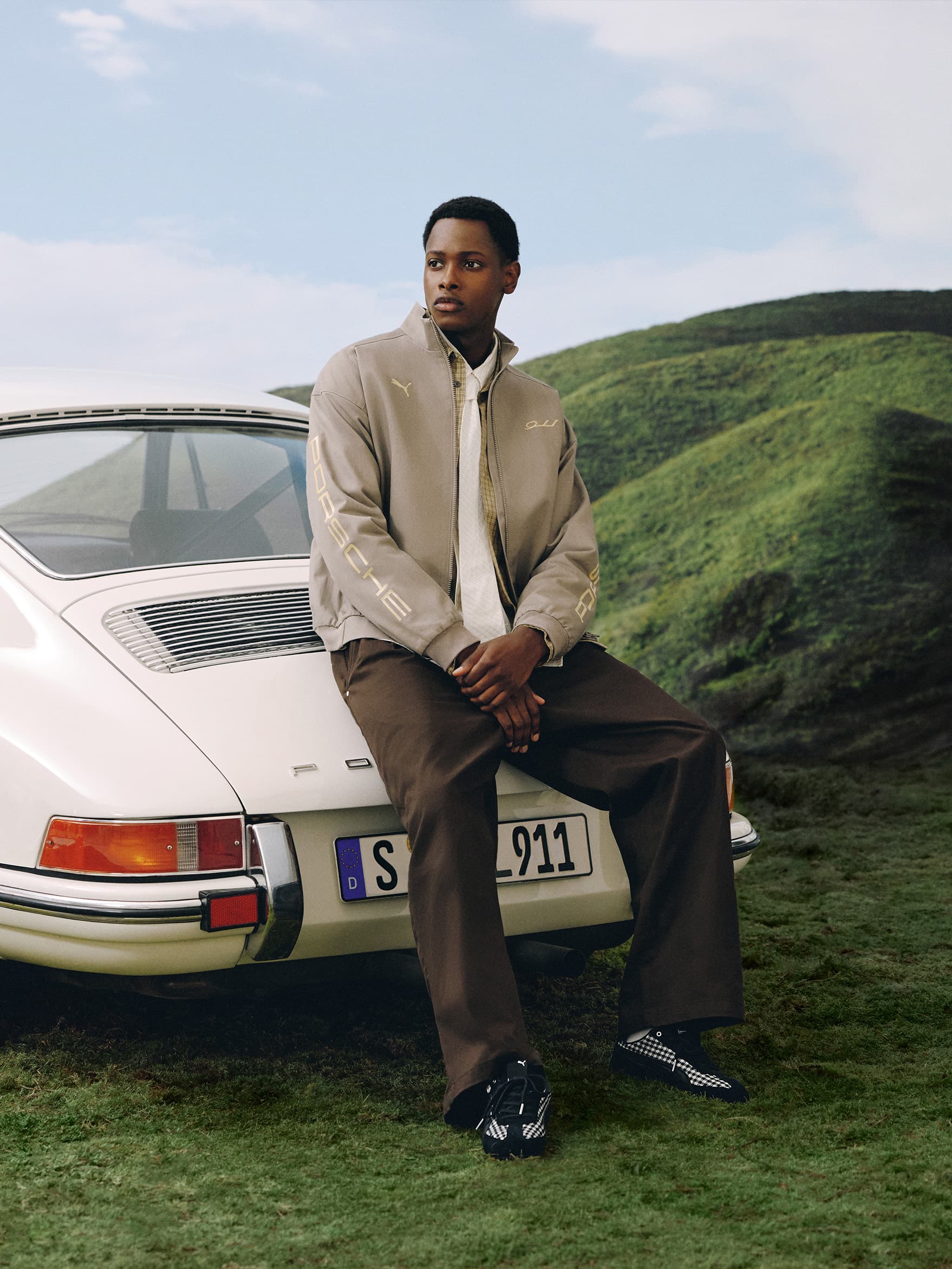 Model in beige Porsche jacket sitting on rear of white Porsche 911, with green hills and cloudy sky behind.
